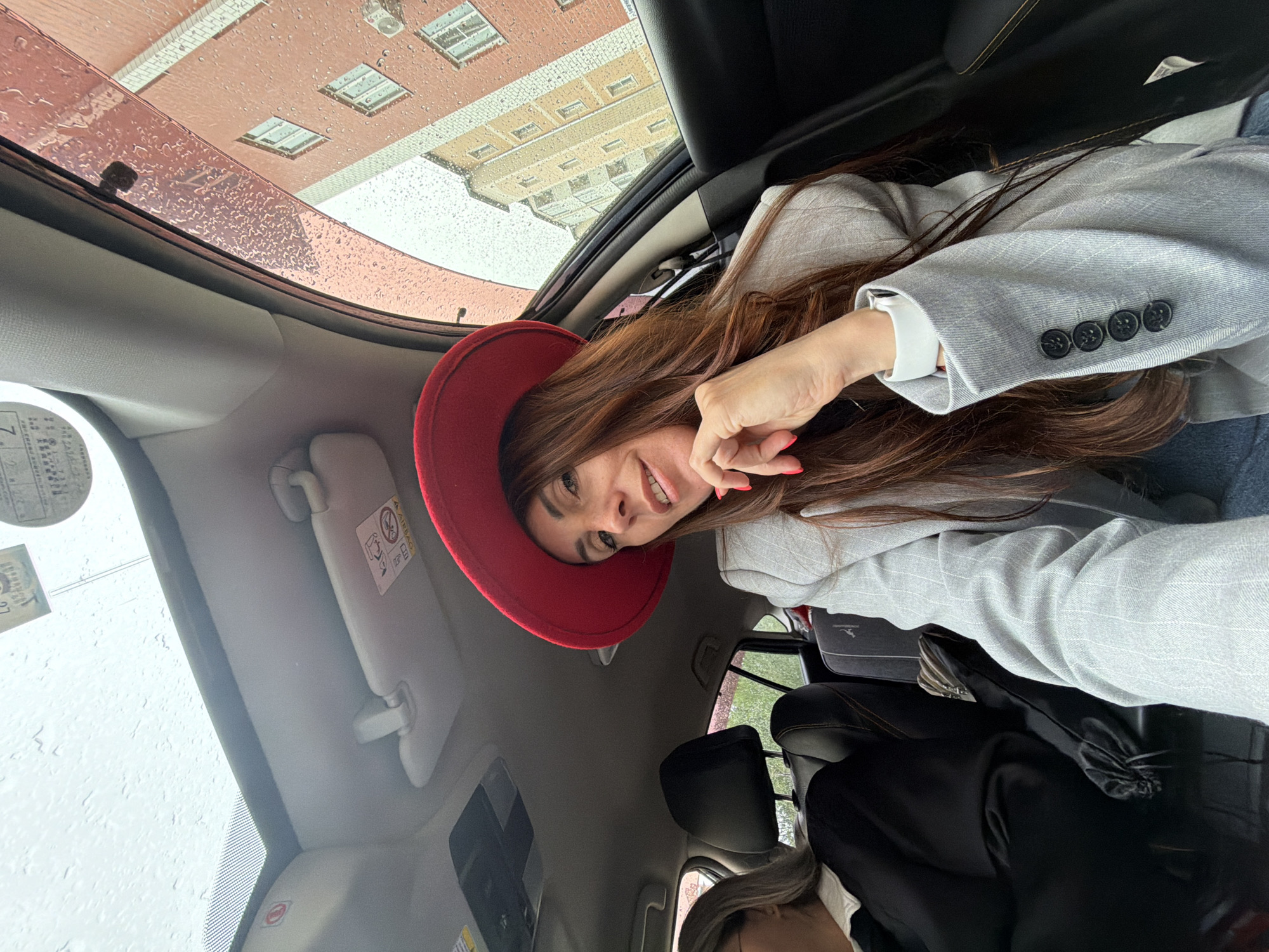 The image captures a woman inside a car, looking up at the camera with a cheerful, open-mouthed smile. She has long, dark brown hair and is wearing a red hat, a light grey pinstriped blazer with dark buttons on the cuff, and a white shirt. A white wristband, possibly a smartwatch or fitness tracker, is visible on her left wrist.
The car's interior is visible, including parts of the dashboard, a raised sun visor, and a black car seat. A portion of another person's head with lighter-colored hair is just visible in the lower right corner. On the sun visor, there is an airbag warning label with a graphic of a child in a car seat and partially legible text, including "AIRBAG". A sticker or permit, featuring numbers and letters like "L6", is affixed to the left side of the windshield.
Through the car's panoramic roof, an exterior scene unfolds. Buildings with red and beige brick facades and numerous windows are visible, suggesting an older urban architectural style. Numerous raindrops are evident on the car's glass surfaces, indicating a rainy or recently rainy day with an overcast sky. The overall lighting suggests it is daytime.
The scene is set in a vehicle, likely in an urban environment, consistent with the provided location context of Birobidzhan, Russia.