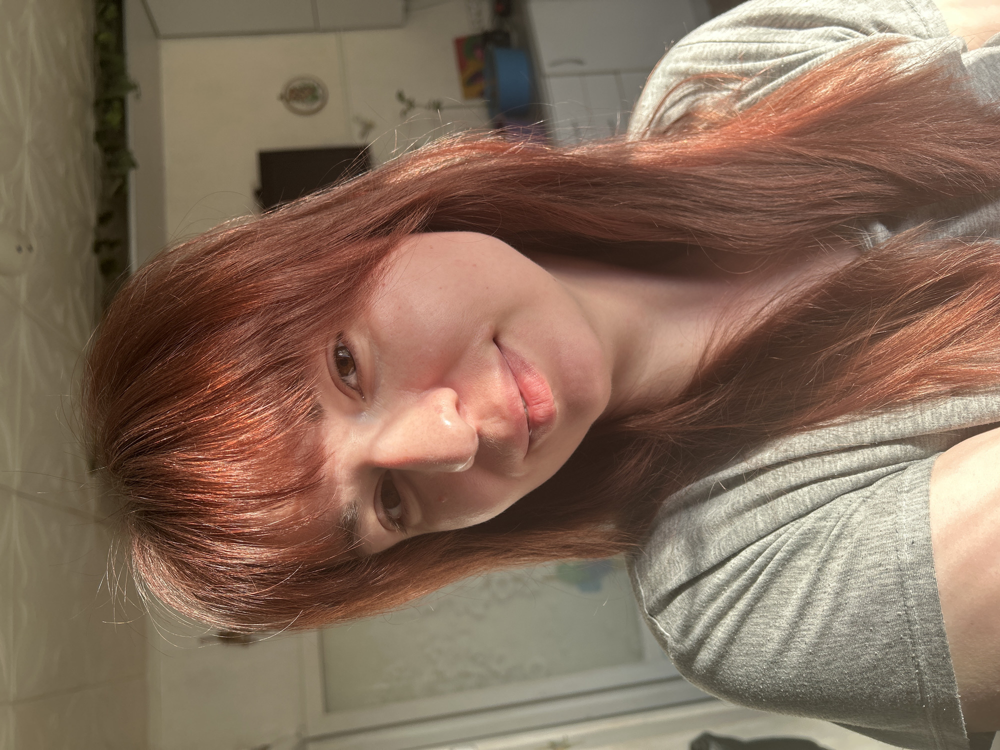A red-haired woman is positioned in the lower left portion of the frame, her face turned towards the camera with a slight smile. She is wearing a gray garment. The background shows a glimpse of an interior space, including a window and what appears to be a kitchen area with cabinets. There's no discernible information to confirm the city is Фоки, Russia.