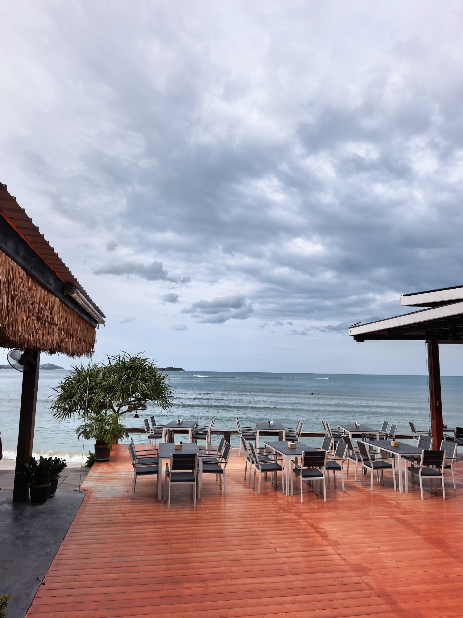 This image captures an outdoor dining area with a view of the ocean in Ko Samui District, Thailand. The foreground features a vibrant, orange-stained wooden deck furnished with numerous tables and chairs. The tables are mostly dark-colored with metal frames, and are set with what appear to be small decorative items. Many of the chairs have a dark seat and backrest with metal legs.
To the left, a thatched roof structure overhangs part of the deck, with a ceiling fan visible beneath it. A spiky green plant in a pot sits near the edge of the deck, and further left, there are more potted plants on a darker section of the flooring.
The middle ground is dominated by the ocean, a calm expanse of blue-grey water meeting a cloudy sky. Gentle waves are visible rolling towards the shore. In the distance, a small island or landmass can be seen on the horizon, and a boat is moving across the water.
The sky above is filled with dramatic, dark grey clouds, suggesting overcast or potentially inclement weather. The overall atmosphere is one of serene, yet slightly somber, coastal dining.