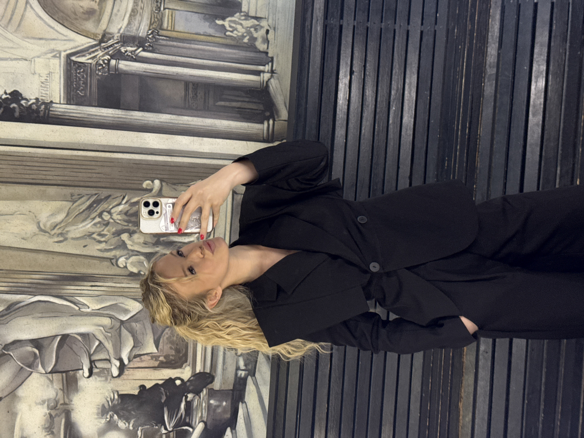 The image shows a young Caucasian woman taking a selfie in what appears to be a museum or art gallery. She is wearing a black blazer and pants, and her blonde hair is styled in loose waves. Her nails are painted red. She is holding a smartphone in her right hand, and her left hand is resting on her hip. The background features a large mural or fresco depicting classical figures and architecture. The lighting is dim, suggesting an indoor setting, possibly in the evening or with limited natural light. The overall atmosphere is elegant and sophisticated, with a hint of playfulness from the selfie. The location context provided suggests this might be in Saint Petersburg, Russia. There is no visible text in the image.