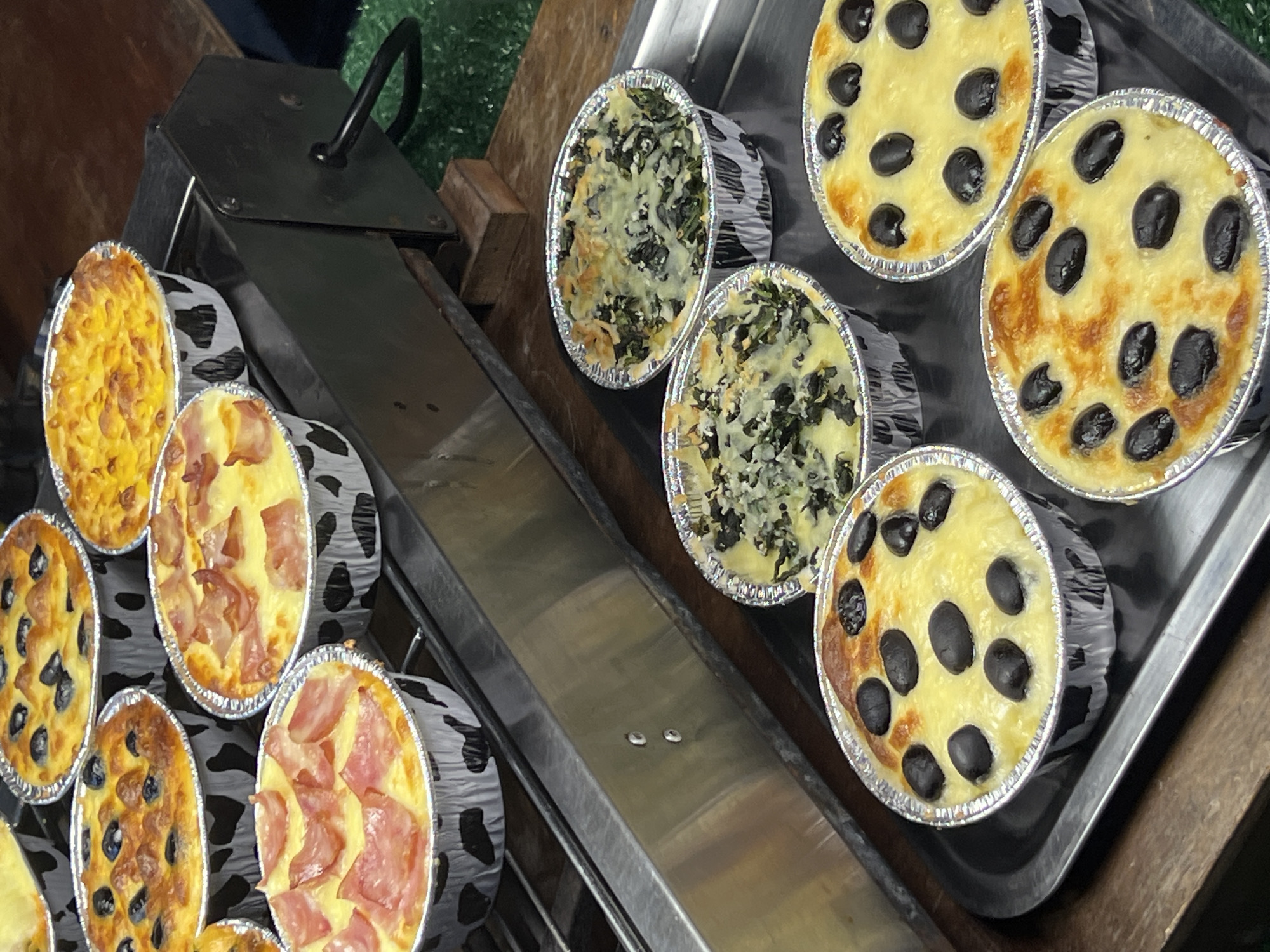 A collection of freshly baked savory pies, or quiches, are displayed in individual aluminum foil containers with a black and white cow print pattern. The pies are arranged on a metal rack, and some are on a larger metal tray.
The pies on the top rack appear to be of two varieties: one filled with a yellow custard and blueberries, and another with a similar yellow base topped with crumbled bacon. The pies on the lower shelf, placed on a metal tray, include a spinach and cheese filling, and a pie topped with black olives and melted cheese.
The background suggests an outdoor or semi-outdoor setting, possibly a market stall or food cart. The surface below the metal rack appears to be a wooden table, and some green material, possibly astroturf or a mat, is visible to the right. The overall impression is of a food vendor preparing or displaying their products. The scene is likely located in the Ko Pha-ngan District of Thailand, given the context provided. No people are visible in the frame.