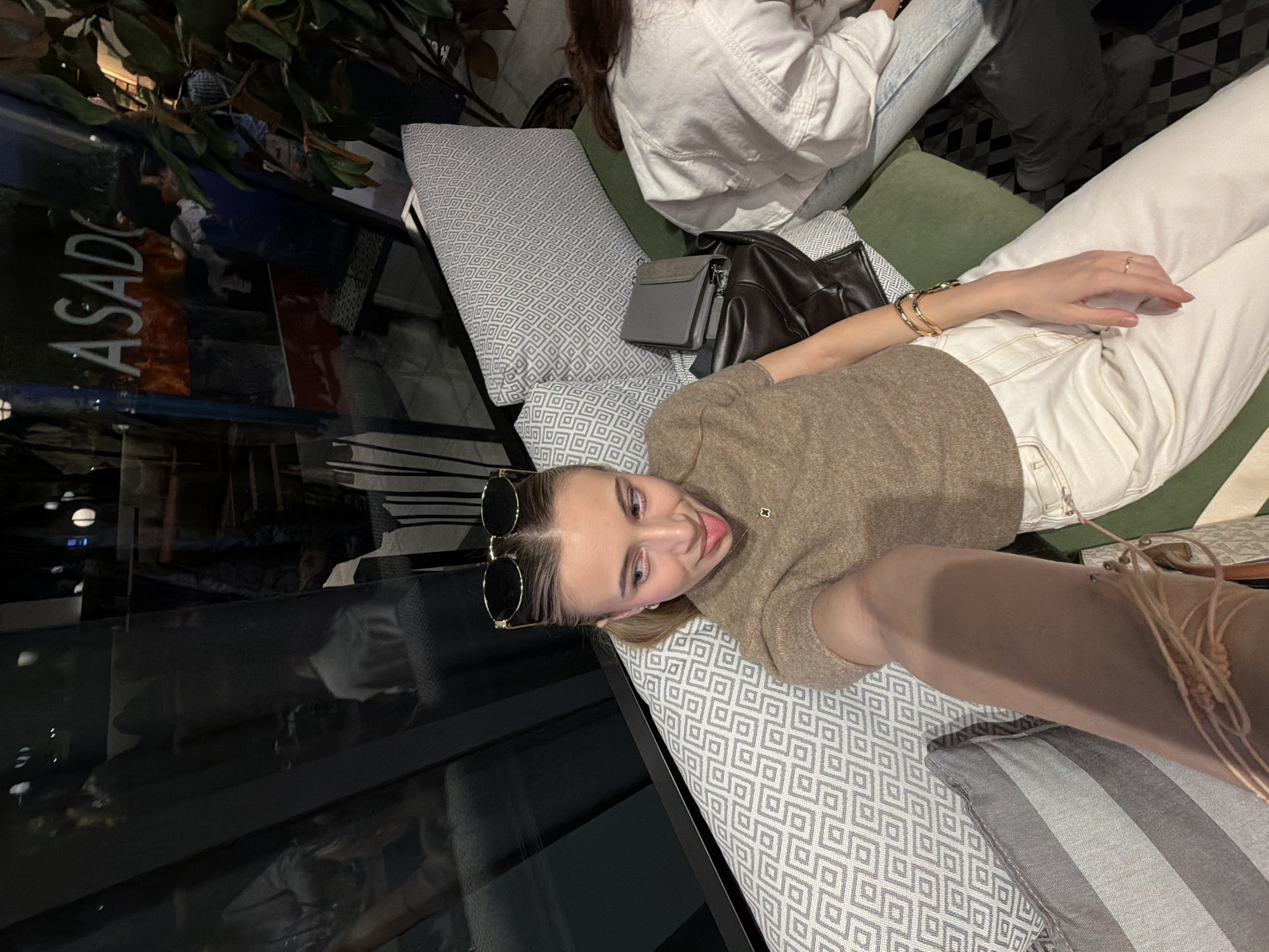 A young woman is seated on a grey and white geometric-patterned couch with a green backrest, taking a selfie. She has light-colored hair in a ponytail, with sunglasses resting on her head. She wears a brown short-sleeved top, white pants, a small pendant necklace, a ring on her right ring finger, and two gold-toned bangles on her right wrist. Her left arm is extended towards the camera. She looks to the viewer's left with her tongue slightly extended. To her right, a second individual is partially visible from behind, wearing a white jacket over light-colored jeans. A grey wallet and a dark leather jacket are placed on the couch between the two. Behind the subjects, a large window reflects the illuminated "ASADO" sign of the ASADO Grill & Wine establishment, confirming the location in Plovdiv, Bulgaria. Blurred reflections of ambient lights and shapes are visible in the window, suggesting an evening or nighttime setting. A plant with dark leaves is partially visible on the far right.