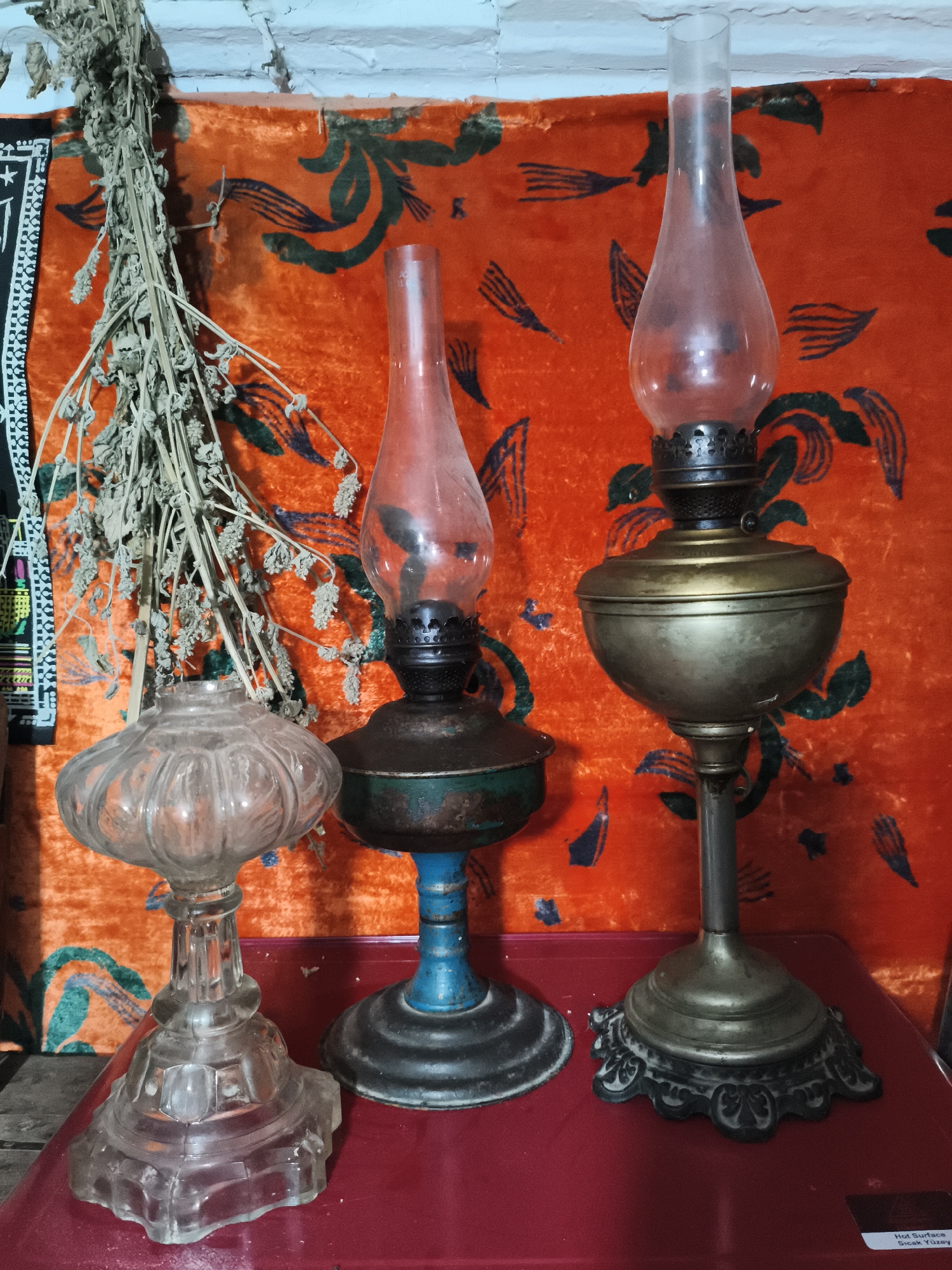 This is a still life photograph taken indoors, likely in Irgali, Russia, showing three oil lamps and a bunch of dried herbs against an orange, patterned fabric backdrop. The lighting is dim, casting soft shadows. The foremost object on the left is a clear, intricately cut glass oil lamp with a bulbous base and a fluted, pumpkin-shaped reservoir. To its right, and slightly behind, stands a brass and dark metal oil lamp with a blue-painted metal stem and a tarnished metal base. On the far right is another brass oil lamp, taller and more ornate than the second, with a wide, flattened reservoir and an elaborate, dark metal base. A bundle of dried, pale brown herbs, suspended from above, hangs to the left of the lamps, partially obscuring the fabric. The fabric features a repeating pattern of dark green, stylized shapes resembling leaves or feathers against a vibrant orange background. The lamps and herbs rest on a flat, red surface, possibly a table. The background wall, visible at the top of the image, appears to be made of rough, white brick or plaster.