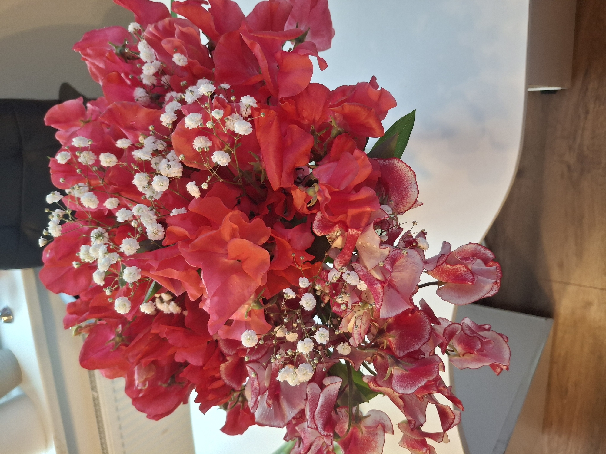A bouquet of sweet peas, primarily in shades of red and pink with some lighter pink and white variations, is displayed against a blurred background. The flowers are interspersed with small white blossoms, likely baby's breath. The bouquet is resting on a white surface, which appears to be a table, with a hint of wooden surface visible below. The lighting suggests an indoor setting. There is no indication of the city of Ogre, Latvia, or any other specific location.