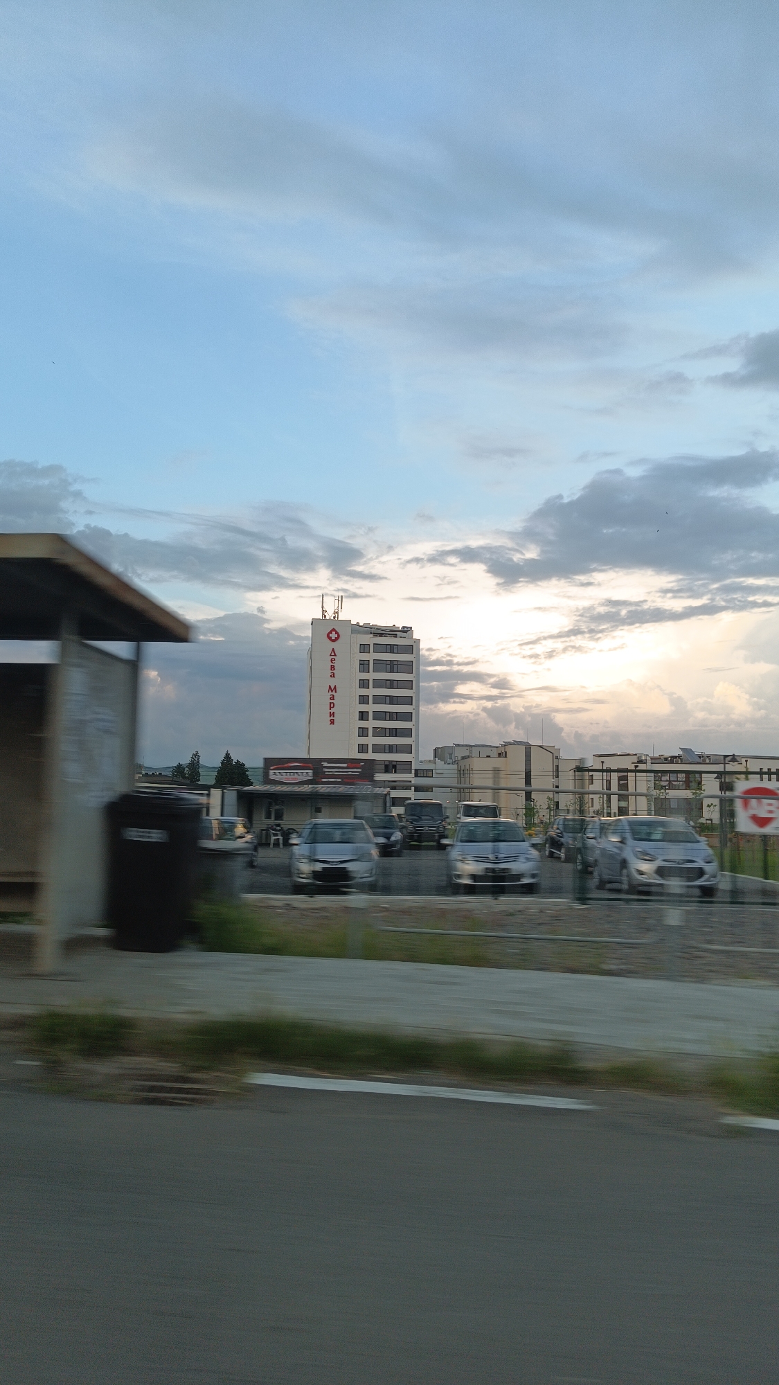 The image depicts a view of the city of Burgas, Bulgaria, with a tall building prominently featuring the name "Дева Мария" in Cyrillic (Deva Maria) on its facade, alongside a red medical cross symbol. The building is located behind a parking area with several silver-colored cars. There is a bus stop on the left side, partially obscured by motion blur, with a trash can visible. The background includes other buildings and a cloudy sky. The photograph seems to have been taken from a moving vehicle, as evidenced by the blurry asphalt road in the foreground.