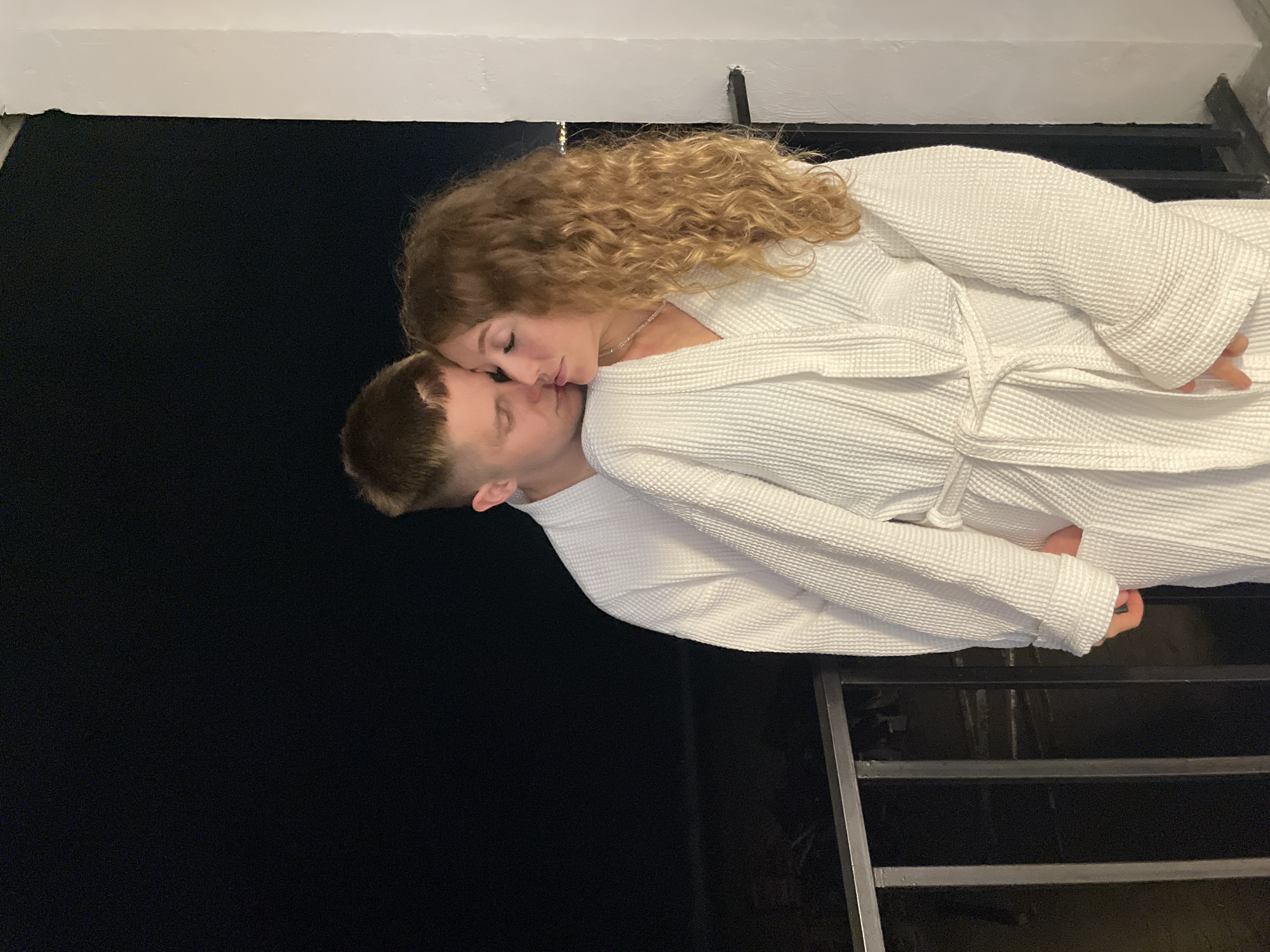 A woman with curly blonde hair and a man are standing close together. They are both wearing white bathrobes. The woman is leaning down to kiss the man on the forehead. They appear to be on a staircase. The location is Unknown, Unknown.