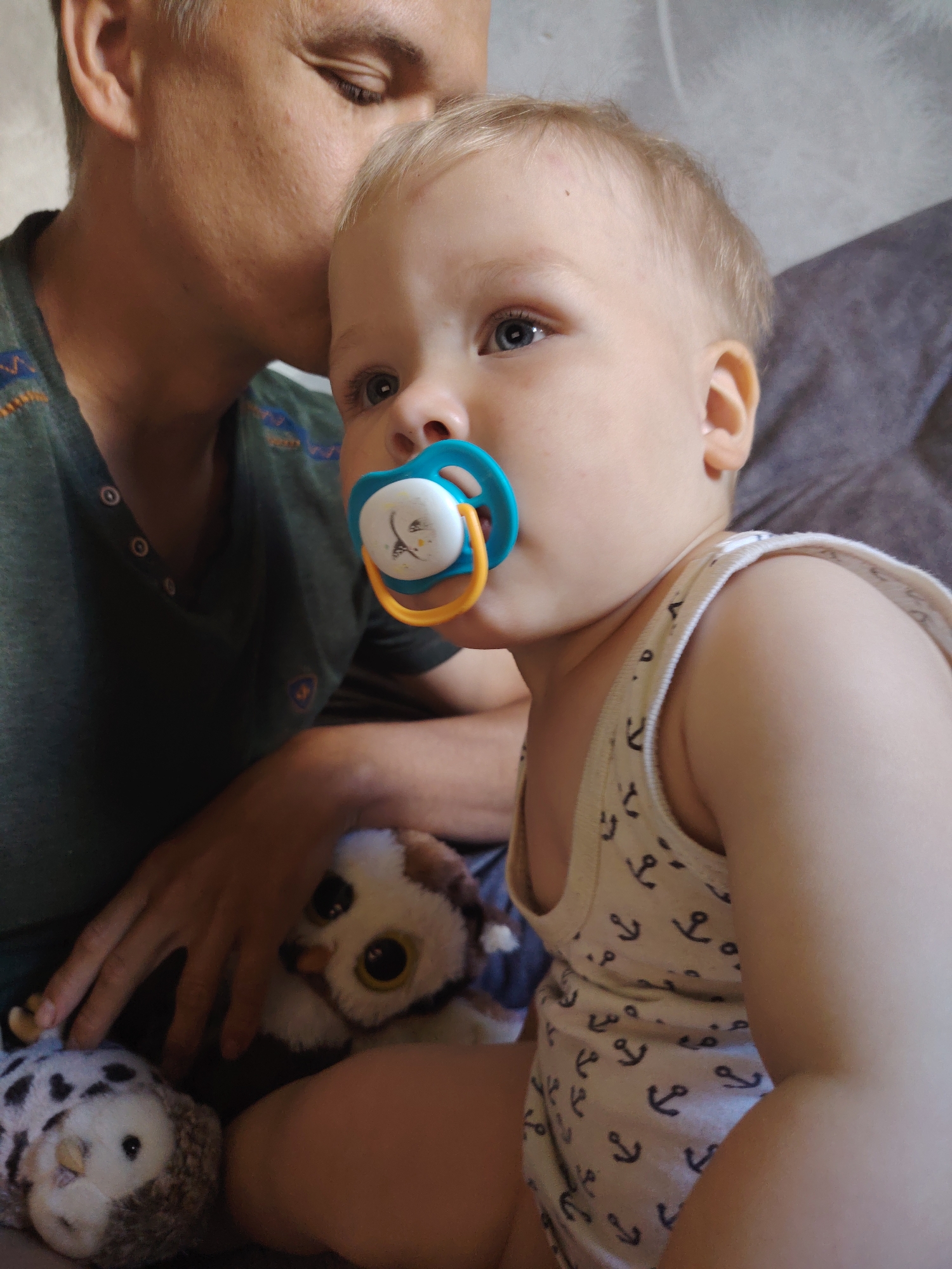 The image shows a close-up of a baby with a pacifier in its mouth, being held by an adult whose face is partially visible. The baby is wearing a white tank top with an anchor pattern. Two stuffed owl toys are visible in the lower left portion of the image. The background appears to be indoors, with a couch or bed and a wall. There are no specific visual cues to indicate the location within the city of Chaikovsky, Russia.