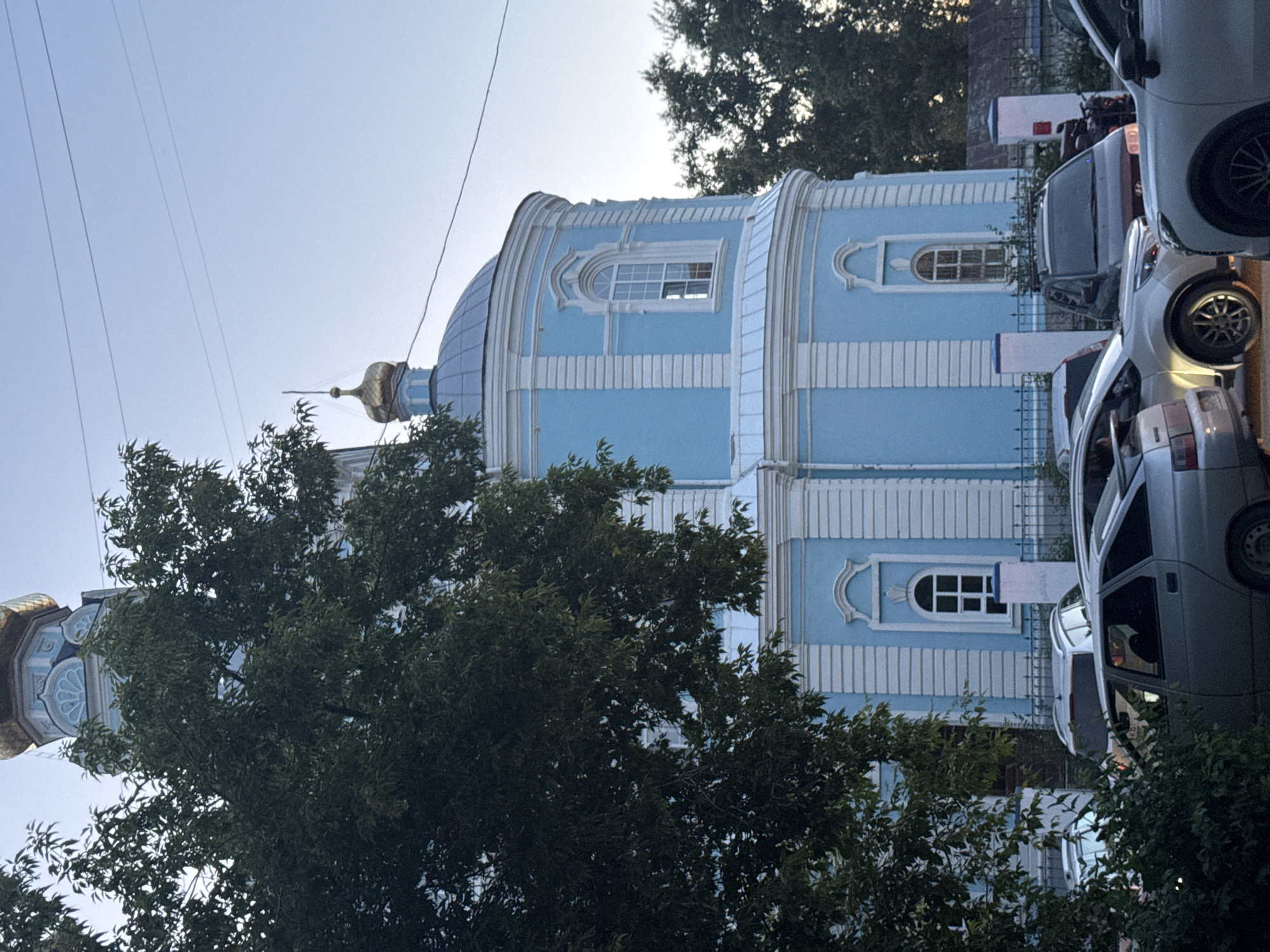 The image shows the Church of the Holy Apostle Andrew the First-Called in Yekaterinburg, Russia. The church is painted a light blue with white trim and features a golden dome. In the foreground, several cars are parked on a street, and a large tree with green leaves partially obscures the view of the church. The sky is clear and light blue.