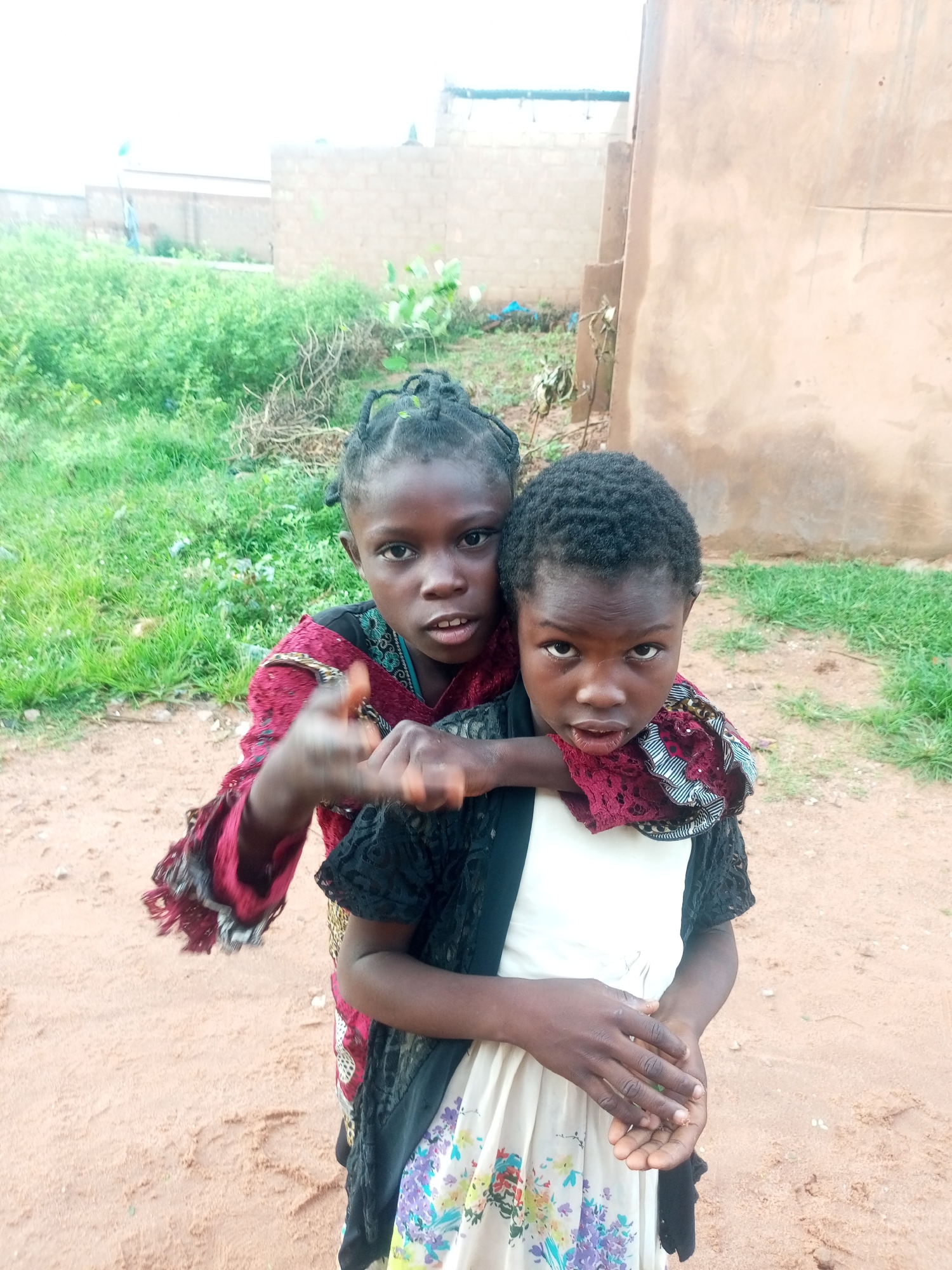 This media file features two young girls in Tempure, Nigeria. The girl in the foreground, on the right, is looking directly at the camera with a neutral expression. She is wearing a white dress with a floral pattern and a black lace overlay. The second girl, positioned behind and slightly to the left of the first, is also looking at the camera. She has her arms around the first girl's shoulders and is wearing a maroon dress with intricate patterns and red and black trim. Her hair is styled in braids.
The setting appears to be outdoors, with a sandy ground and sparse vegetation in the background. A light-colored, unfinished building is visible behind the girls. The lighting suggests it could be daytime, possibly overcast. The overall scene conveys a candid moment between the two children. There is no visible text in the image.