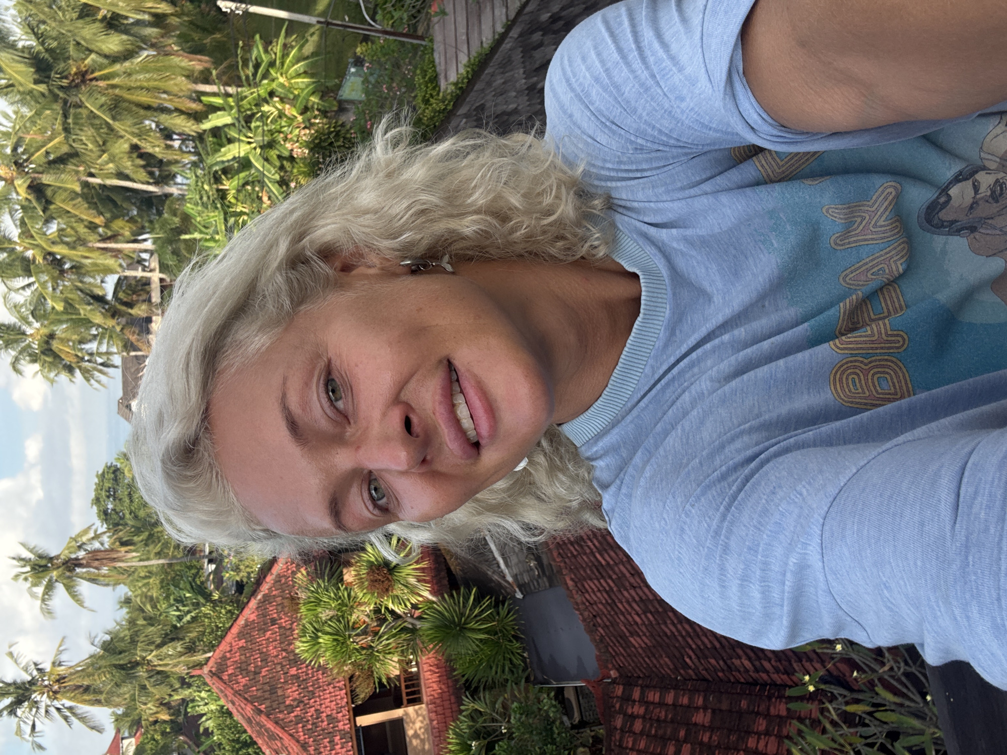 A medium shot captures a fair-skinned, fair-haired woman, smiling slightly. She is wearing a light blue t-shirt with a vintage-style graphic, against a lush tropical backdrop. The location appears to be Unknown, Unknown, with dense greenery including palm trees and tropical foliage visible in the background. The red-tiled roofs of traditional buildings are also visible, suggesting a residential or resort area. The image is taken from a slightly elevated perspective, giving a view of the surrounding landscape.