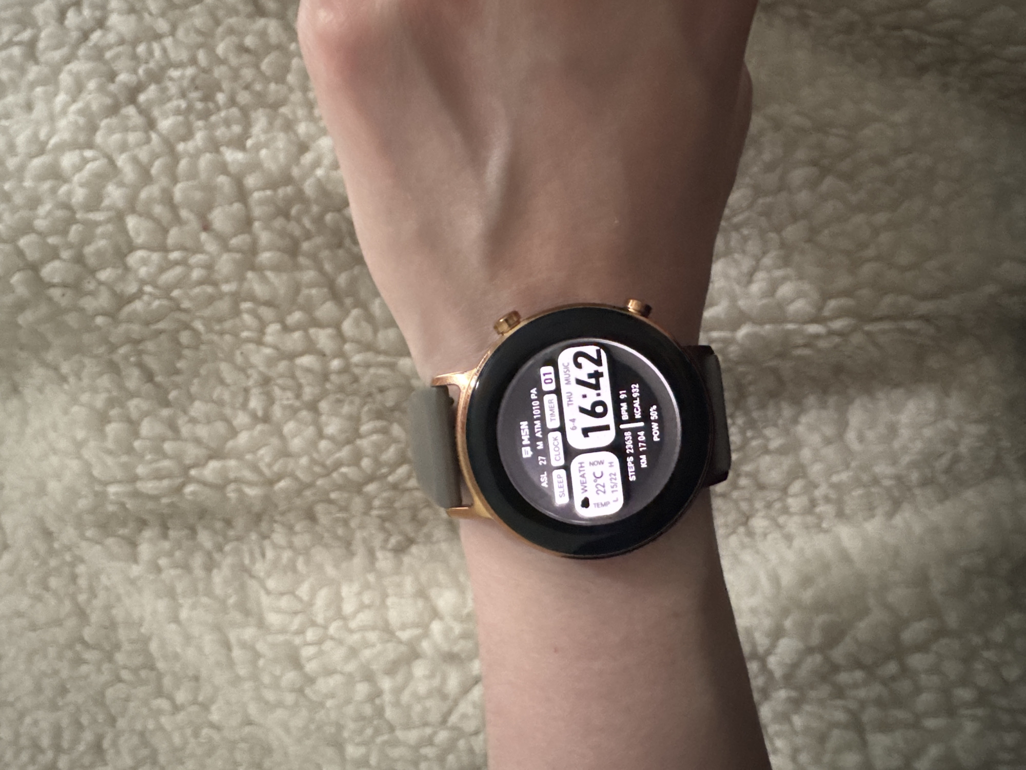 A person's left arm, adorned with a smartwatch, rests on a light-colored, textured surface resembling a fluffy blanket. The smartwatch features a round display with a rose gold or copper-toned bezel and a greyish-brown strap.
The smartwatch screen is illuminated, displaying detailed information. The current time is prominently shown as 16:42. The date reads "6-4 THU," indicating June 4th, Thursday. Weather details include a current temperature of 22°C, with daily low and high temperatures of 15°C and 22°C, respectively. Atmospheric pressure is noted as "ATM 1010 PA," along with "MSN ASL 27 M." This weather data is consistent with the provided location context of Riga, Latvia.
Activity tracking metrics are visible, including "STEPS 23638," "KM 17.04," "KCAL 932," and "BPM 91." The watch's battery level is at "POW 50%." Shortcuts or widgets for "SLEEP," "CLOCK," "TIMER," and "MUSIC" are also displayed on the watch face. The scene appears to be an indoor setting, with no particular activity taking place beyond wearing and displaying the smartwatch.