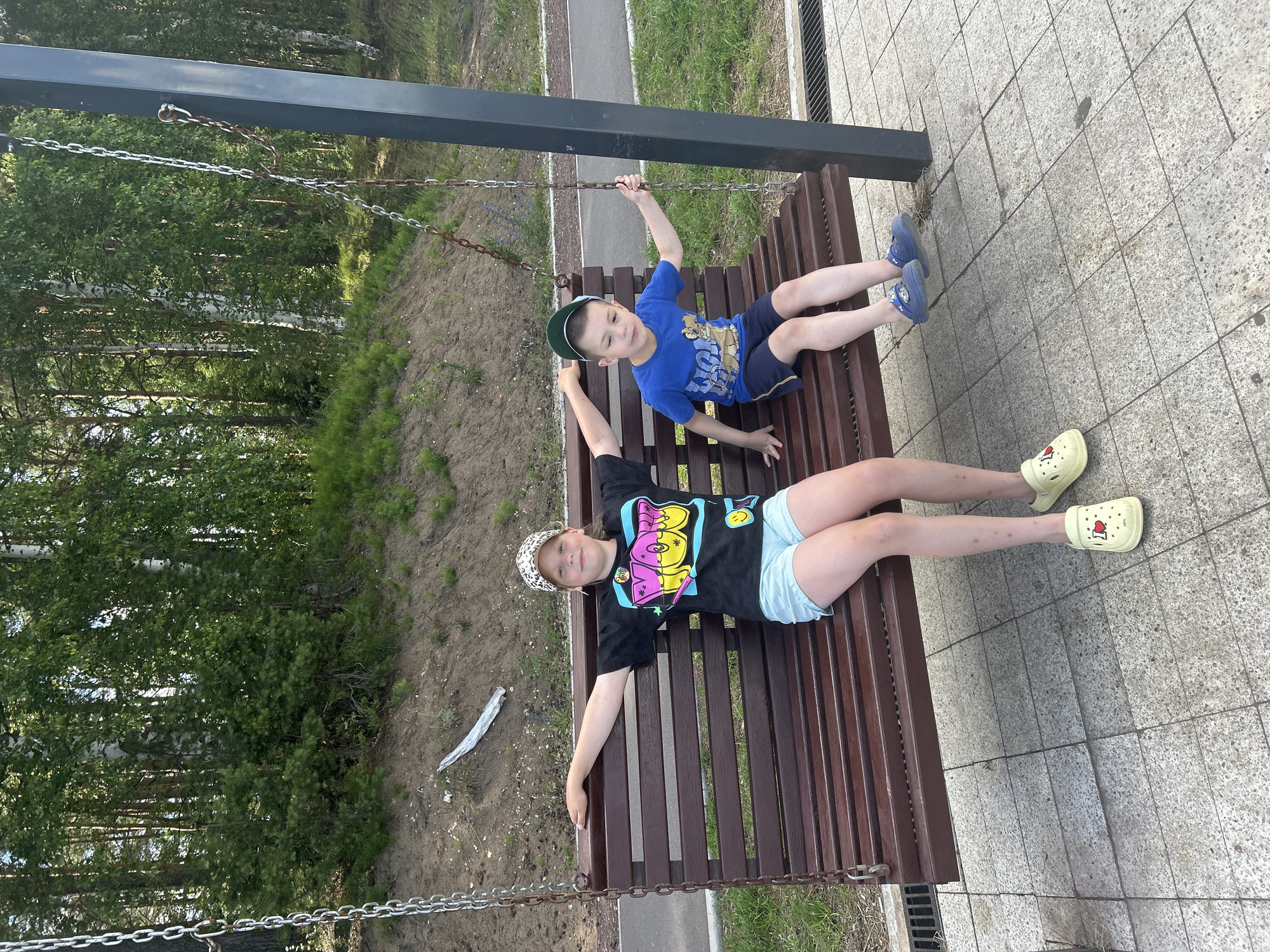 Two children, a girl and a boy, are sitting on a wooden swing in a park in Chaikovsky, Russia. The girl is wearing a black t-shirt with a colorful graphic, light blue shorts, and a patterned baseball cap. The boy is wearing a blue t-shirt with a graphic and navy blue shorts, and a green baseball cap. They are both facing forward, and the girl has her arms outstretched. The swing is suspended by chains from a dark metal post. The background features trees and a paved path. The ground is covered with concrete tiles and some grass.