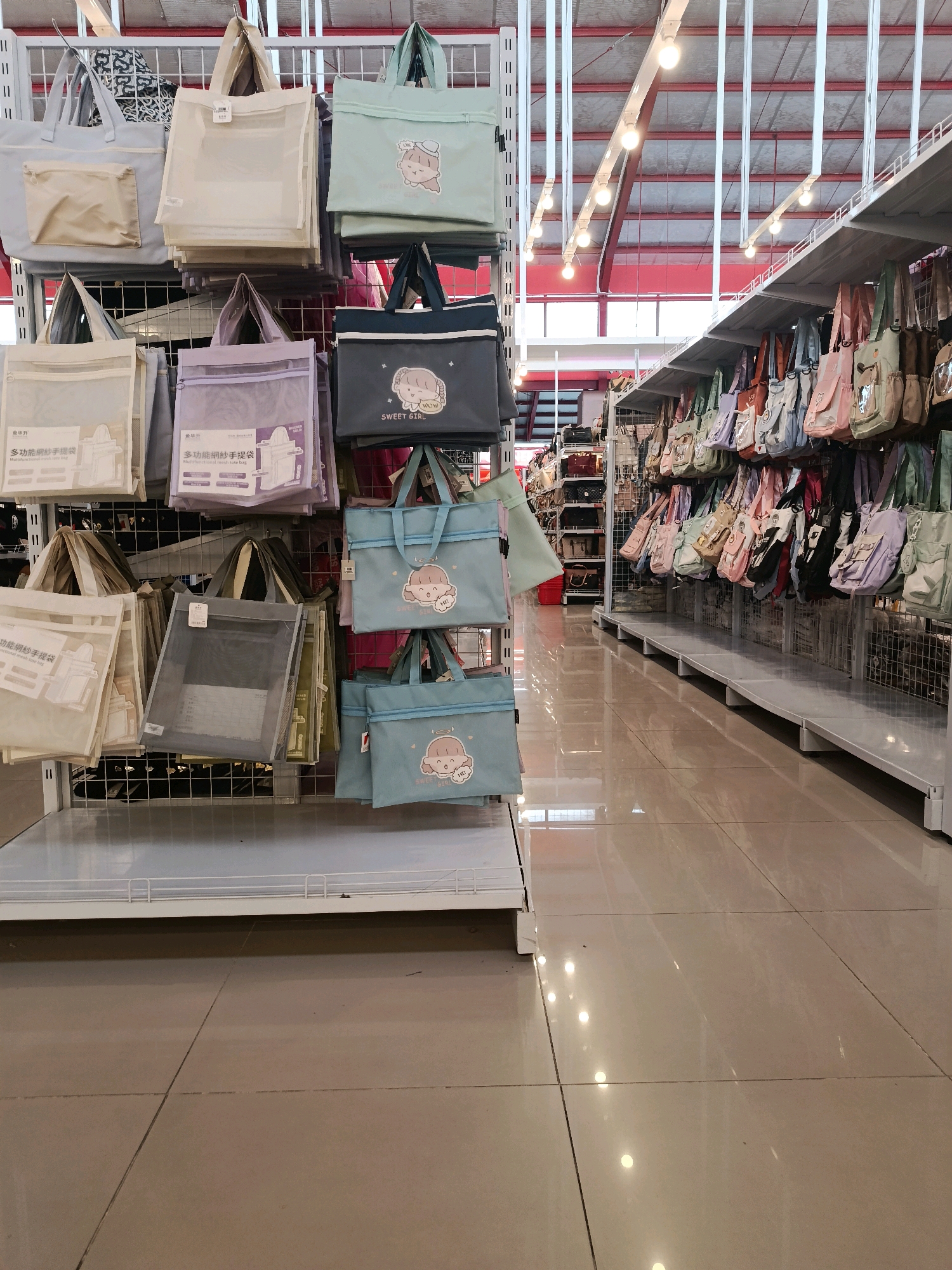 The image depicts a retail store interior, likely a section dedicated to bags and accessories, with rows of shelves displaying various tote bags. The bags are predominantly in pastel colors such as light blue, mint green, cream, and lavender, and many feature cartoonish illustrations of animals or figures, often with text like "SWEET GIRL". They are hung on metal racks and displayed in staggered rows, suggesting a retail environment. The store has a bright, modern aesthetic with white shelving units and a tiled floor that reflects the overhead lighting. In the background, more shelves filled with a wider variety of bags can be seen stretching into the distance. The overall impression is one of a well-stocked and organized shop. The specific location in Bulukumba, Indonesia, is not discernible from the visual information provided in the image.