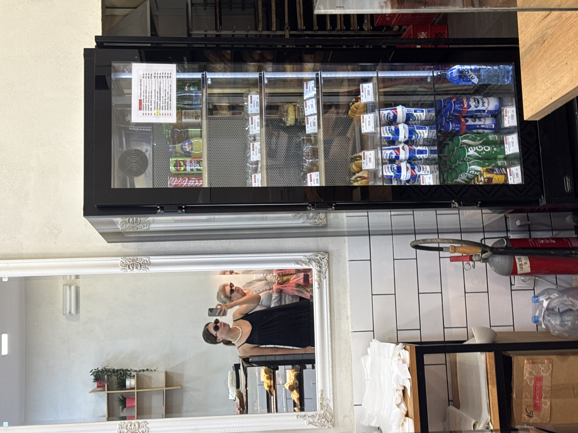 The image shows the interior of a cafe or bakery in Belgrade, Serbia. In the foreground, a large refrigerated display case is filled with various drinks and food items, including yogurts, juices, sodas, sandwiches, and desserts. A price list is visible on one of the shelves. To the left of the display case, there is a mirror reflecting two women. One woman in the mirror is wearing a black dress and sunglasses, and is holding a smartphone to take a selfie. The other woman is older and wearing a cardigan, holding a shopping bag. The background of the reflection shows shelves with baked goods. To the left of the mirror, a wooden shelf unit with potted plants is visible. In the lower left corner, there are stacks of white paper bags and a cardboard box. A red fire extinguisher is positioned next to a white tiled wall.