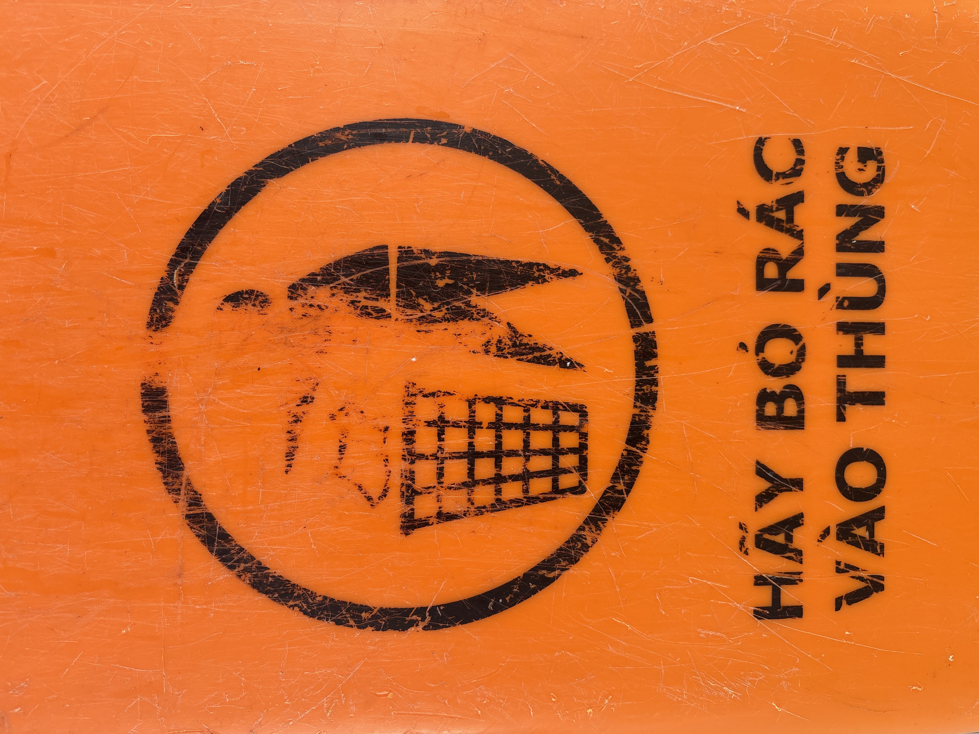The image shows a close-up of an orange surface with black text and a circular graphic. The graphic depicts a stylized person throwing trash into a bin. The text below reads "HÃY BỎ RÁC VÀO THÙNG," which translates to "Please put trash in the bin." The orange surface appears to be made of plastic and is scratched and worn, suggesting it is an outdoor sign or label. The location is unknown.