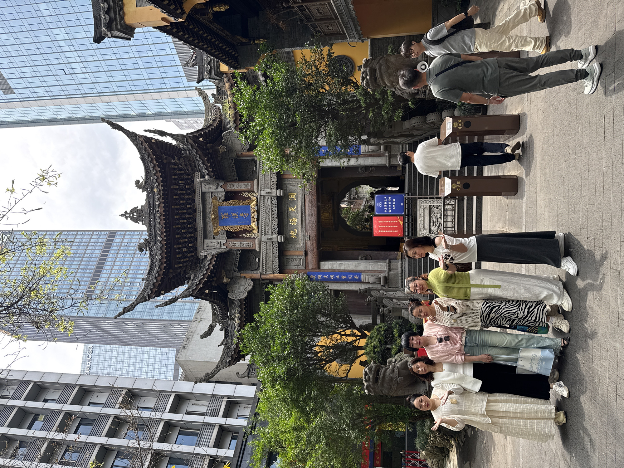 The image captures a group of six women standing in a row on a paved area in front of the Arhat Temple (Luohan Si) in the Yuzhong District of Chongqing, China. The temple, recognizable by its traditional Chinese architectural design and intricate carvings, serves as the backdrop for the group. The women are dressed in various styles, some wearing lighter colors and traditional attire, while others sport more modern clothing.
Behind the temple, towering modern skyscrapers contrast with the ancient architecture. To the right, individuals pass by the entrance to the temple, while a tree frames part of the temple's structure. Signage in Chinese is visible near the entrance, along with stone lion sculptures flanking the temple's entrance. The lighting indicates a sunny day with the image taken in daylight conditions.