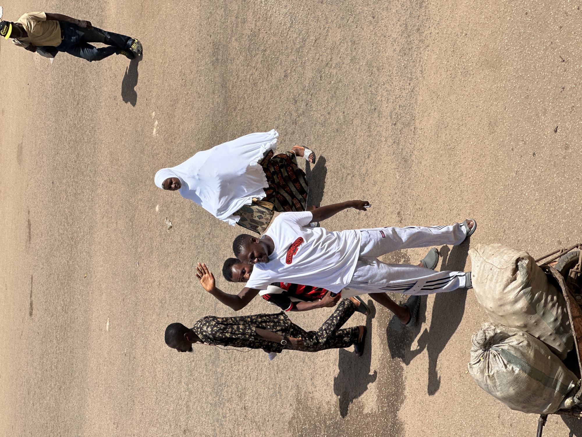 In Bukuru, Nigeria, a group of people are walking on a dusty, paved surface under bright sunlight. In the foreground, two large burlap sacks are visible, some contents spilling from one, resting in a trolley. To the right and slightly behind the sacks, a man wearing a white t-shirt with a red logo and grey sweatpants with black stripes is waving with his right hand. He has a person in a red and black checkered shirt partially visible behind him. Further back and to the left, a man in a patterned black and gold shirt with black pants and headphones is walking. To the right of the man in the white t-shirt, a woman is wearing a white hijab and a patterned skirt. In the background, a man in a tan shirt and blue jeans, wearing a yellow cap, walks away from the camera, carrying a dark bag over his shoulder. The environment appears to be an outdoor public space with scattered debris on the ground.