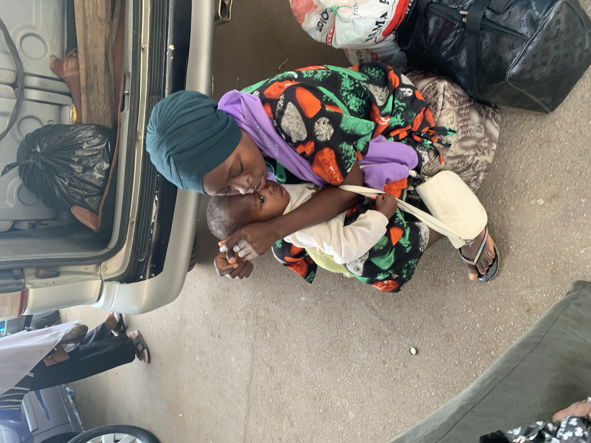 A woman wearing a dark green head wrap, a purple sash, and a multicolored dress holds a baby in a white shirt. They are kneeling on a concrete surface. A white bag, a black patterned bag, and a bag with red and white text are to the right. Part of a silver vehicle is on the left. The location is inferred to be Bukuru, Nigeria.