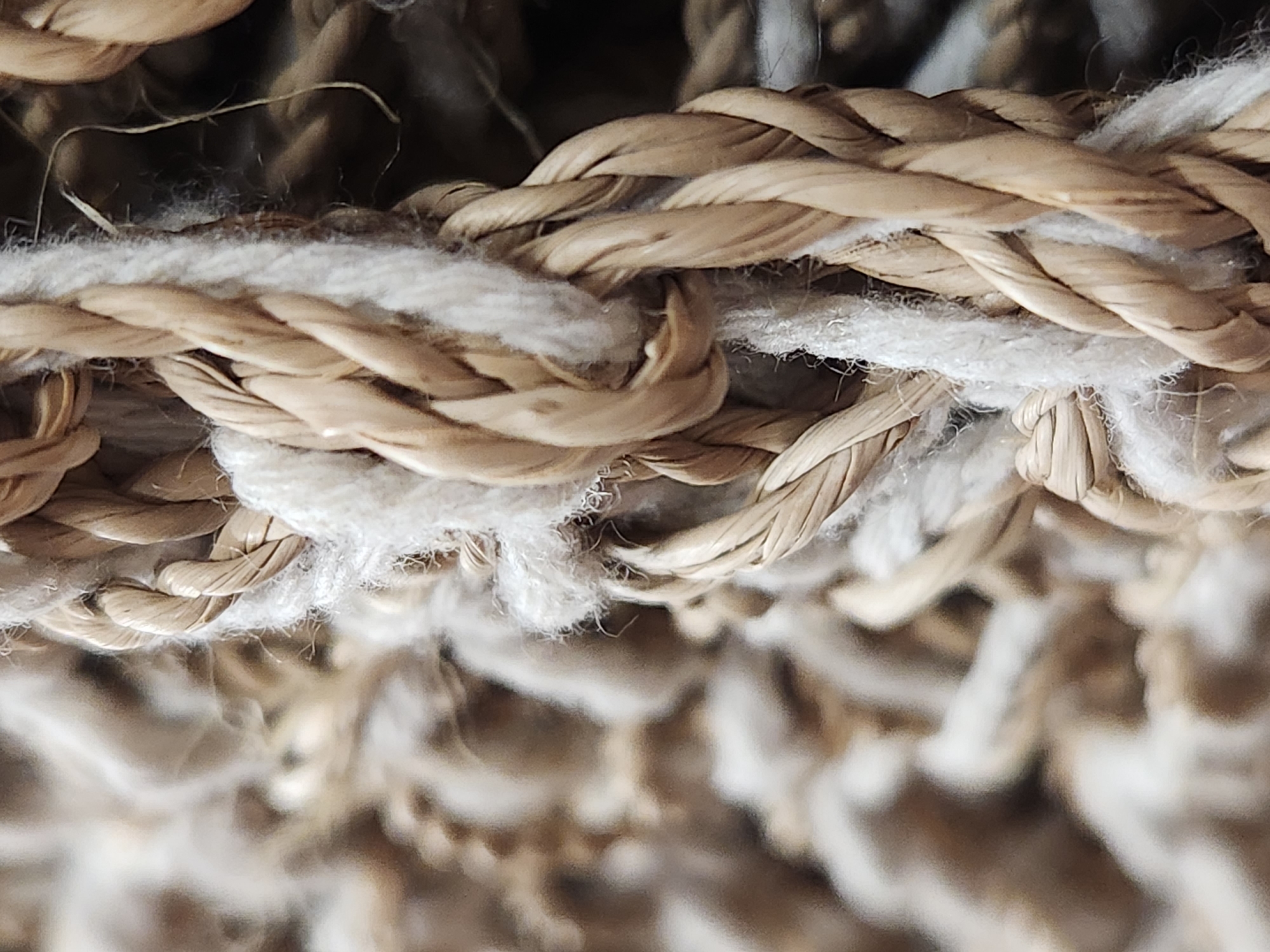 The image is a close-up, macro photograph of thick, braided ropes. The ropes appear to be made of natural fibers, with varying shades of beige and brown. Some of the ropes are tightly woven, while others are looser and more frayed, with white fibers interspersed. The background is out of focus, showing a blurry texture of similar materials. There are no people, animals, or discernable signs of a city or specific location.