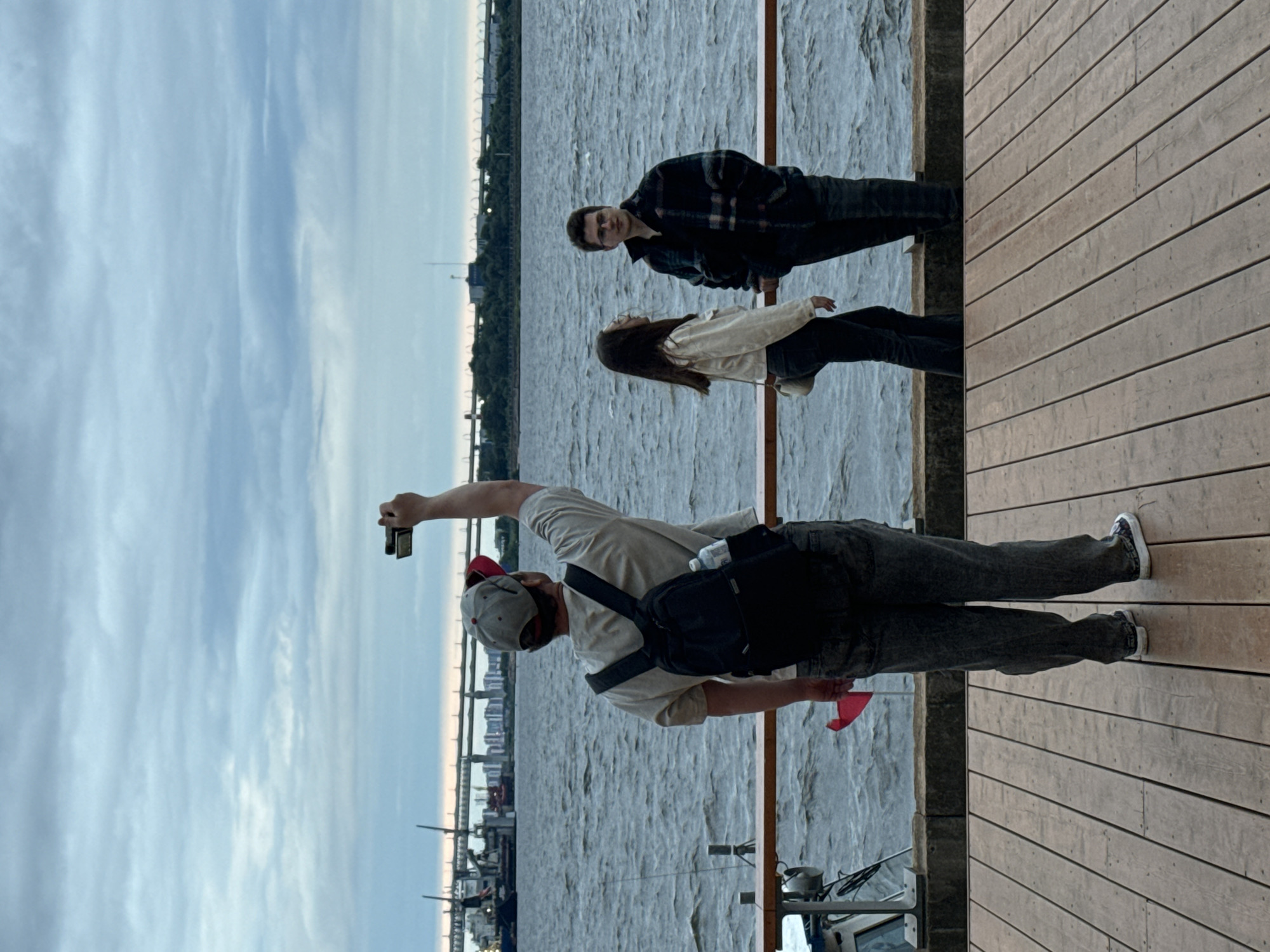 The image depicts a view of the Neva River in Saint Petersburg, Russia, on an overcast day. In the foreground, a person wearing a baseball cap, t-shirt, and jeans stands on a wooden deck, holding a phone up to take a photo or video. They have a backpack on and a red flag is tucked into their back pocket. To the right, a young woman in a cream-colored jacket and jeans stands beside a young man in a plaid jacket and jeans. They are both looking out at the river, with a railing separating them from the water. In the background, a wide body of water, likely the Neva River, stretches towards a distant cityscape with a bridge and industrial structures visible under a cloudy sky. The water is choppy, indicating wind.