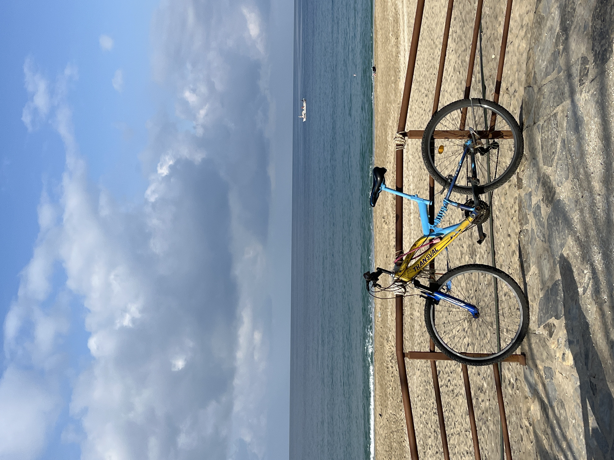A bicycle is leaning against a metal railing next to a concrete wall. In the background is the sea, a ship, and a cloudy sky. The scene is located in Alanya, Turkey.