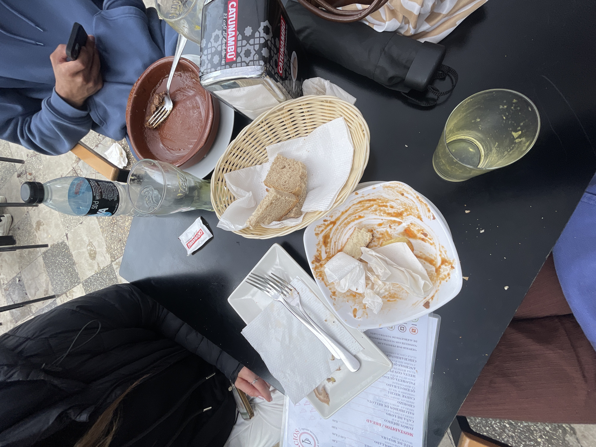 A high-angle shot reveals a table covered with remnants of a meal. A person in a blue hoodie holds a phone on the left side of the table, near a brown bowl and a water bottle. A "CATUNAMBU El café de Andalucía" container sits next to a wicker basket holding pieces of bread and white napkins. A plate smeared with sauce and food scraps, also containing crumpled napkins, sits next to a square dish with cutlery and a menu listing food items and prices. A near-empty glass and a handbag are situated towards the right of the table. A person in a black jacket is visible on the lower-left side. The setting appears to be a restaurant in Jerez De La Frontera, Spain, based on the menu.
