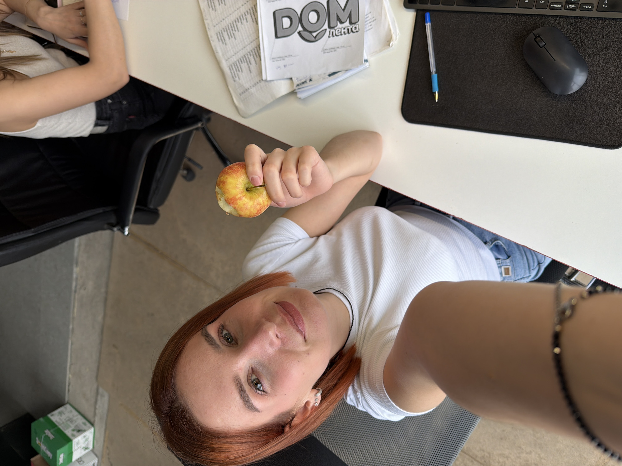 The image captures a female individual in the foreground, taking a selfie in what appears to be an office or workspace environment in Chaikovsky, Russia. She possesses shoulder-length, reddish-brown hair and green eyes, wearing a white ribbed t-shirt, light blue jeans, small silver hoop earrings, and a dark beaded bracelet on her left wrist. Her right hand holds a partially eaten red and yellow apple. Behind her is the back of a dark office chair. To her right, a second female individual is partially visible, seated in a black, high-backed office chair, wearing a white ribbed tank top and dark jeans. Her right arm rests on a light-colored desk. On this desk, a stack of documents includes a publication titled "ДОМ Недвижимость" (DOM Real Estate) in Russian. Also on the desk are a black mouse pad, a black wireless computer mouse, a blue ballpoint pen, and the edge of a black keyboard. The background on the left side of the frame reveals a concrete floor and two stacked cardboard boxes, one green (partially labeled "CKS") and one white.