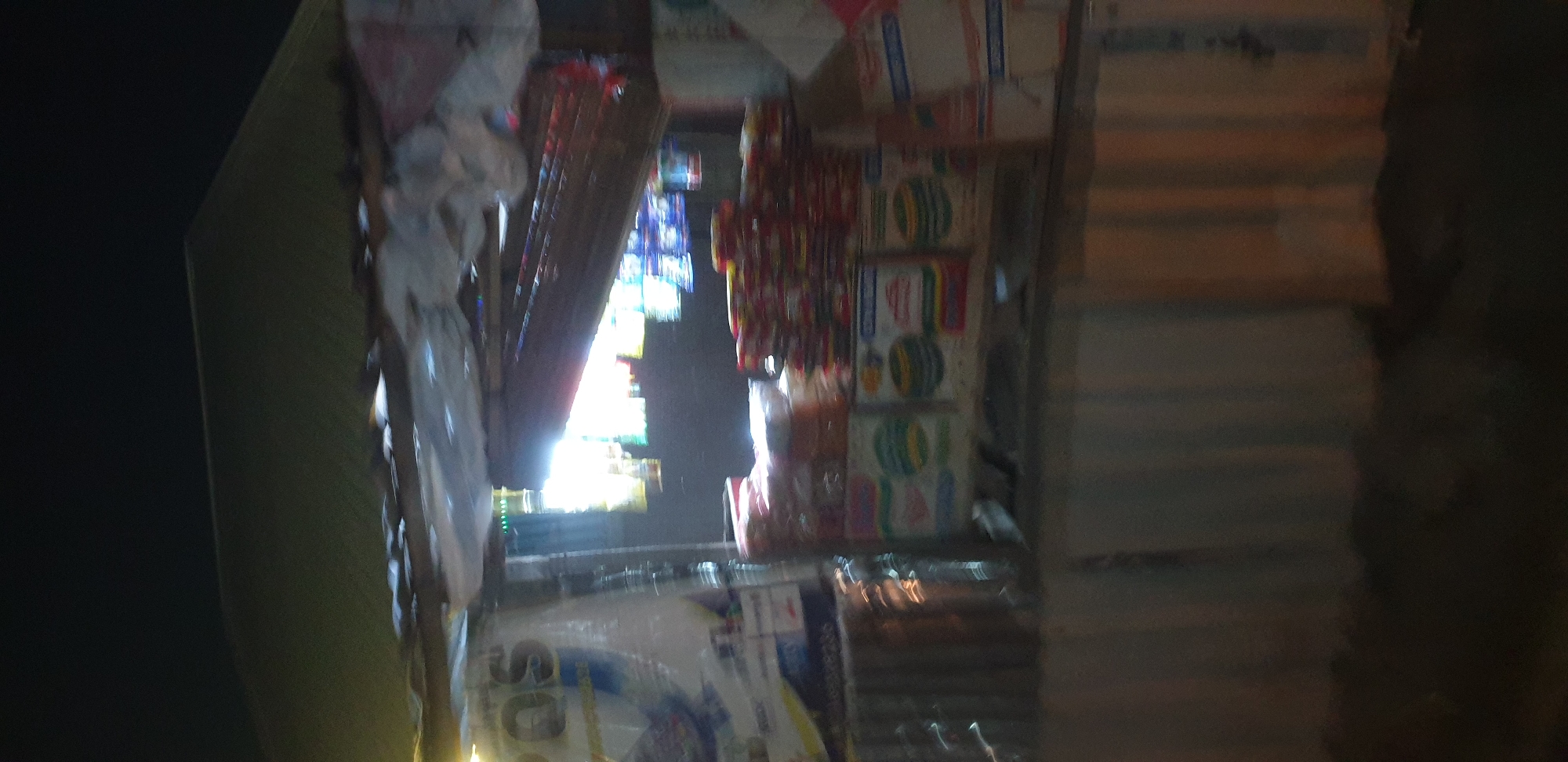 The image depicts a night scene of a roadside stall in Narayi, Nigeria, likely a small grocery or convenience store. The stall is covered with a corrugated metal roof and partially enclosed with corrugated metal sheeting on the sides. The interior of the stall is illuminated by artificial light, casting a bright glow and creating shadows.
Various products are visible on display, including what appear to be packaged food items like cereals, snacks, and possibly canned goods, stacked on shelves and surfaces. Some items are wrapped in plastic or cardboard packaging with visible labels and branding, although the text is largely illegible due to the image quality and distance. A large white plastic bag with partial text, possibly advertising a brand or product, is also visible on the left side of the stall.
The overall environment suggests a typical street scene in a commercial area at night, with the stall serving as a point of sale for everyday goods. The lighting and the arrangement of goods indicate that the stall is open for business. The image quality is low, with significant blur and noise, making it difficult to discern fine details or identify specific products. There are no people clearly visible in the image, and no specific actions or interactions are depicted. The background is dark, consistent with a nighttime setting, with some hints of the surrounding urban environment or natural darkness.
