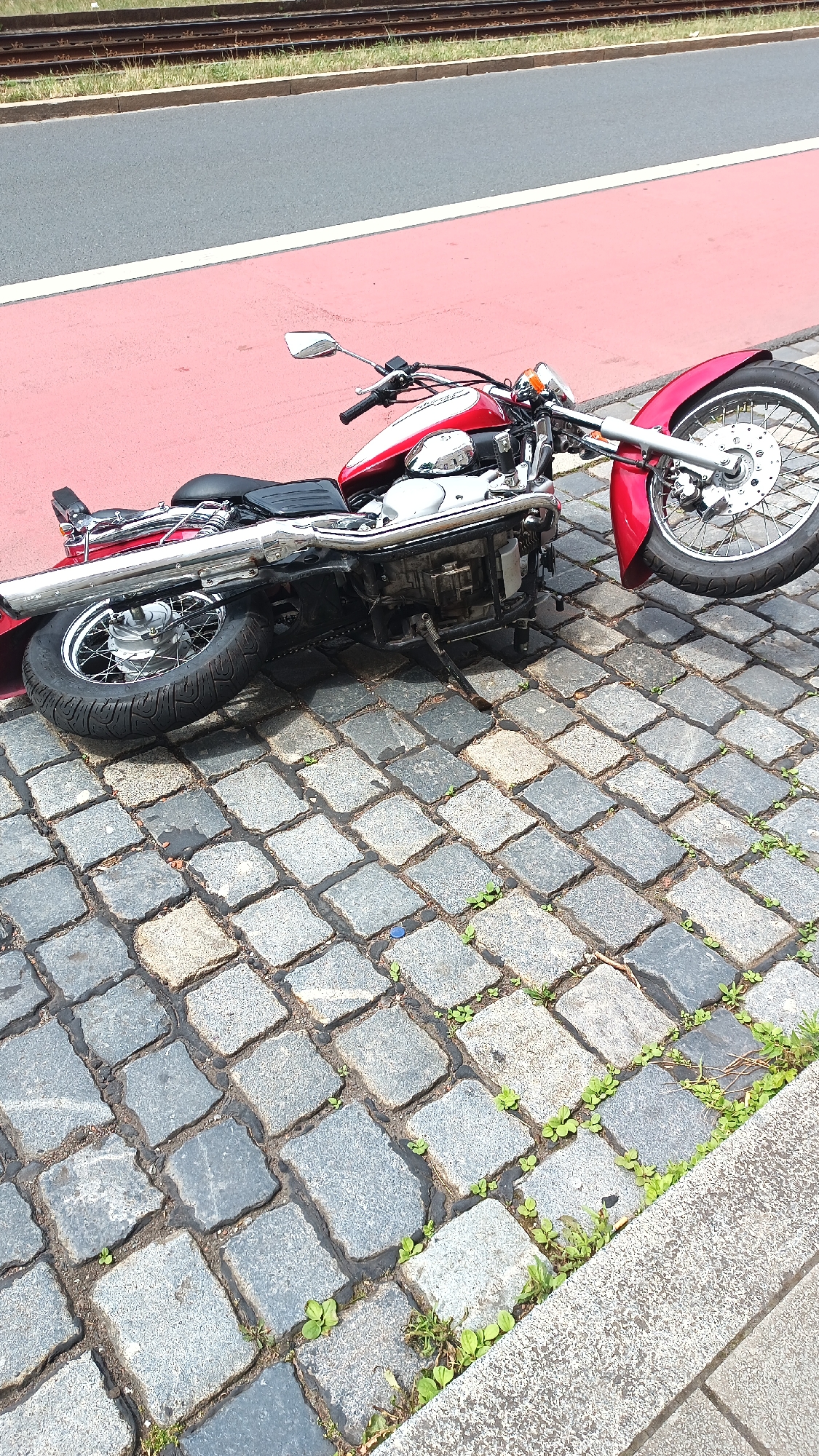 A red and chrome motorcycle lies on its side on a cobblestone street. The motorcycle is a cruiser-style bike with a prominent exhaust pipe and spoked wheels. It appears to be damaged, with the handlebars angled awkwardly and the front wheel resting on the ground. The motorcycle is positioned in the foreground of the image. Behind the motorcycle, there is a red-colored bike lane, and beyond that, a grey asphalt road. In the background, a set of train tracks runs horizontally across the frame, bordered by green grass. The scene is illuminated by natural light, suggesting it is daytime. The cobblestones are wet, and there are small patches of green vegetation growing between them, indicating recent rain or a damp environment. No people are visible in the image. The overall impression is one of an accident or incident involving the motorcycle, with the fallen vehicle resting on the street. The location appears to be urban, given the presence of roads, bike lanes, and train tracks.