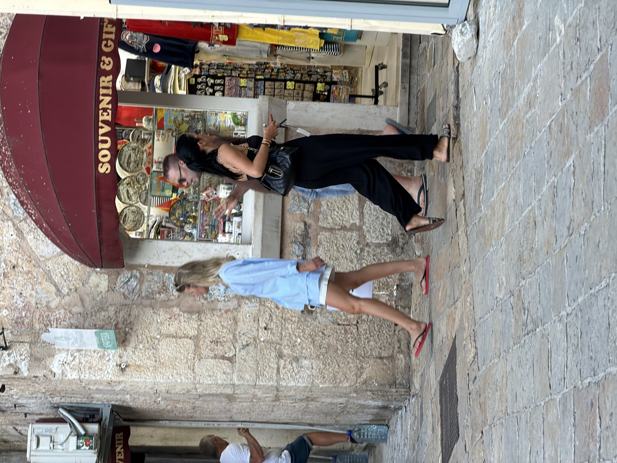 The image depicts a street scene in Budva, Montenegro, with a focus on a souvenir shop. A cobblestone street runs diagonally across the foreground. In the middle ground, three individuals are visible. A woman with blonde hair, wearing a light blue shirt and white shorts, walks from left to right. To her right, a man with sunglasses, wearing a light-colored t-shirt, walks alongside a woman with dark hair, wearing a black strapless top and black wide-leg pants, carrying a black purse. Behind them, a souvenir and gift shop with a red awning is visible. The shop's window displays various items, including plates, flags, and clothing. To the far left, a man in a white t-shirt and blue shorts is partially visible near a wall with an air conditioning unit and a sign. The architecture features rough stone walls and a paved walkway.