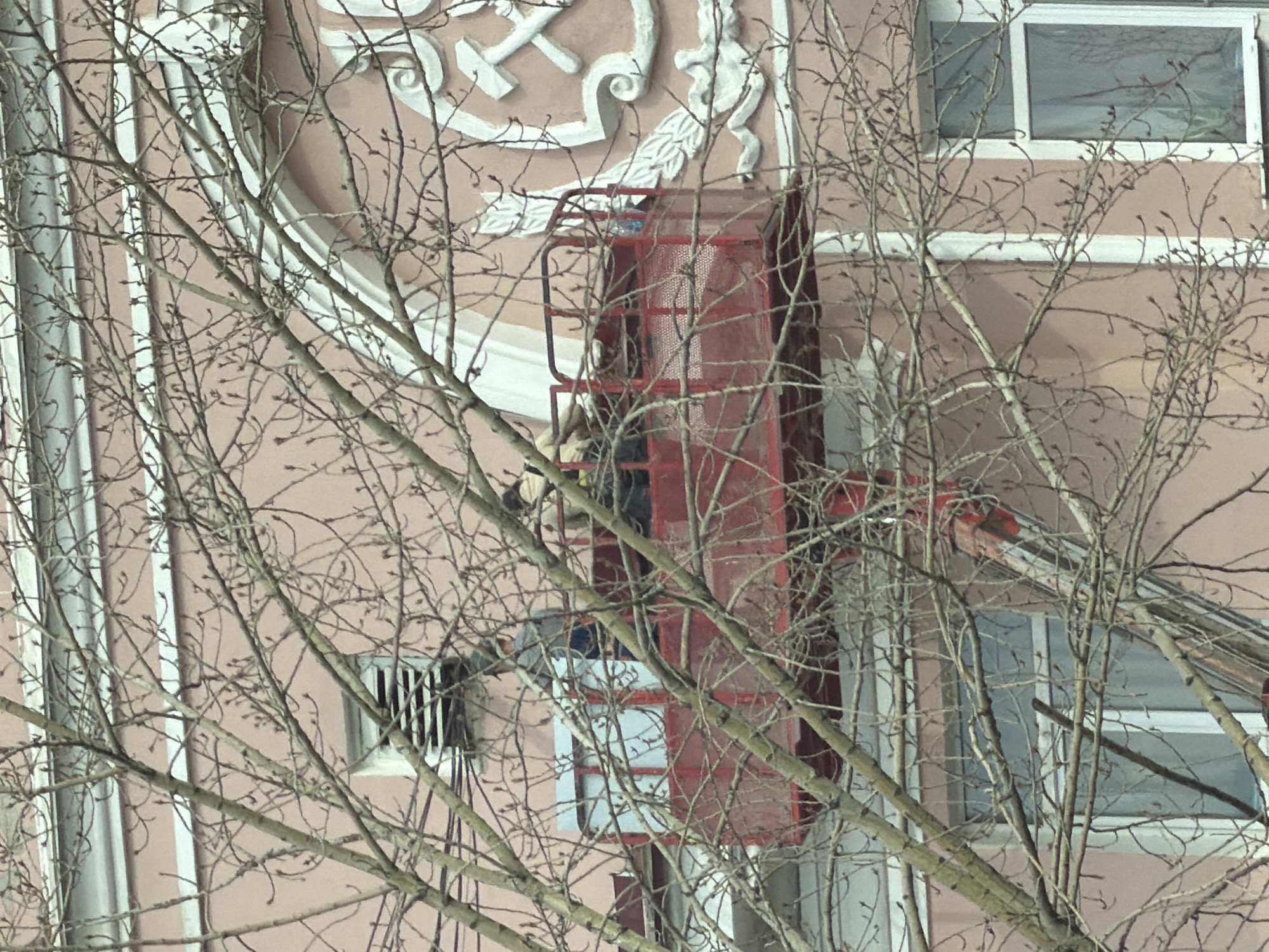 A partially obscured view through leafless tree branches reveals a section of a light pink multi-story building in Perm, Russia. An elevated red service platform with a worker is positioned against the building's facade, likely for maintenance or repair. The building displays decorative white architectural elements on its exterior, including a hammer and sickle symbol and flourishes. The building has multiple windows with white frames, some of which are visible, while others are obscured by the platform and tree branches.