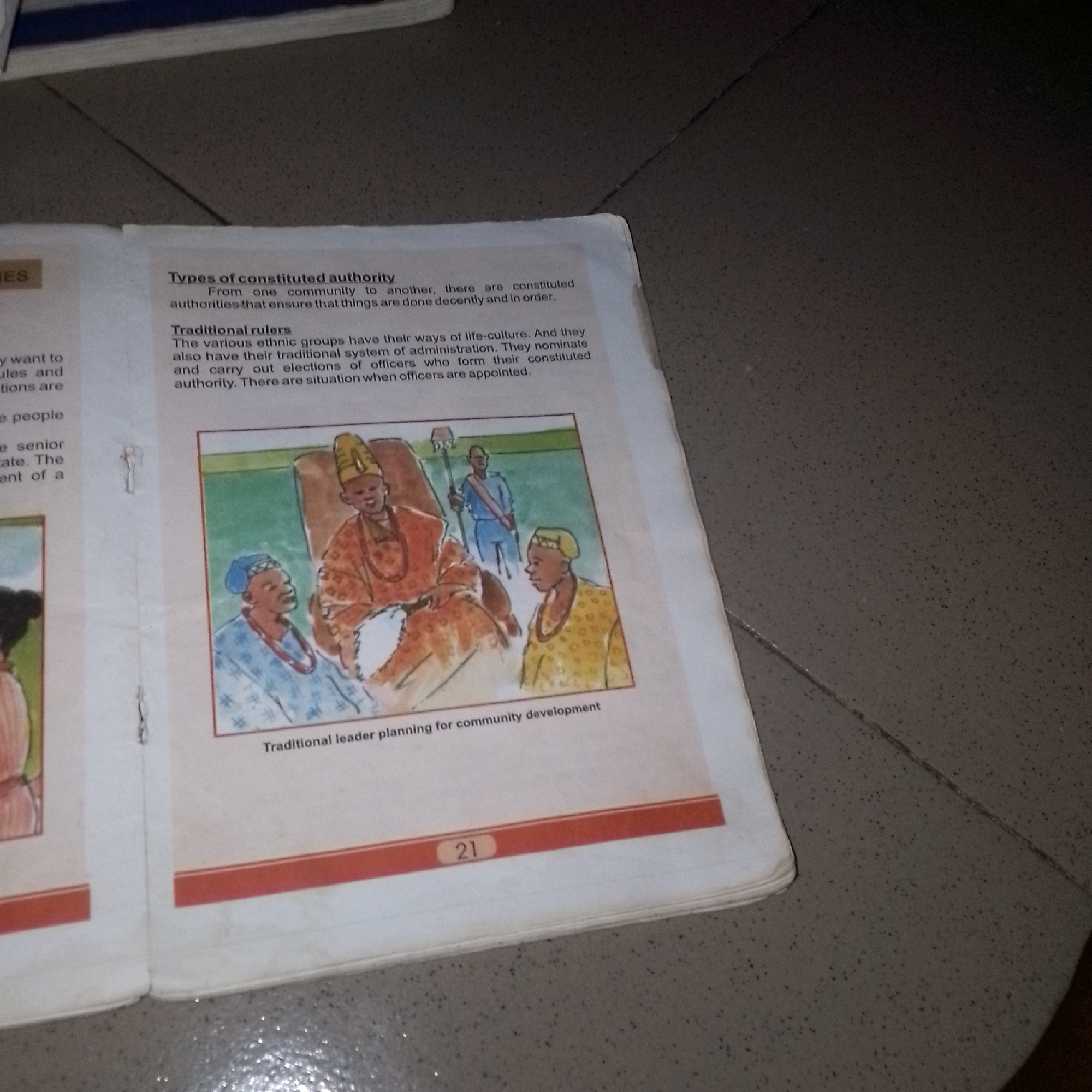 The image shows an open book with text and an illustration. The text is on the right page and discusses "Types of constituted authority" and "Traditional rulers." The illustration depicts a group of people in traditional attire, likely a leader and advisors, engaged in what appears to be a meeting or ceremony. The illustration is in a colorful, somewhat stylized manner. A page number "21" is visible at the bottom of the right page. The book is placed on a textured surface, possibly a floor or table, with a speckled pattern. There is no visible indication of the city of Jalingo, Nigeria, within the image content itself. The image appears to be taken from an elevated angle, looking down at the book.