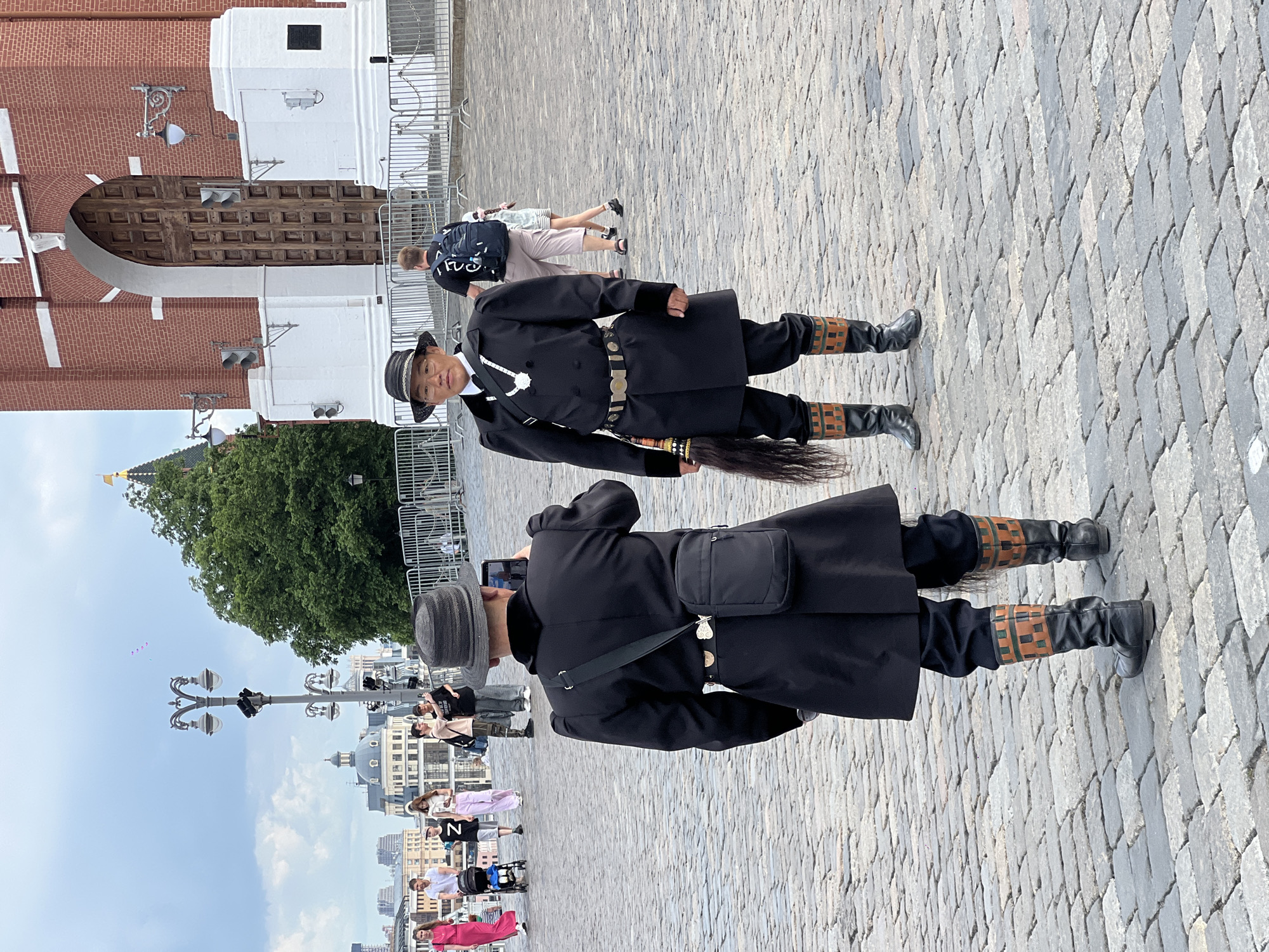 Two men are standing on a cobblestone surface. Both men are wearing dark clothing, including boots, and hats. The man in the foreground is taking a picture with a smartphone. Behind them is a low metal fence with a building and a green tree in the background. In the background is an array of other people, including a pink-dressed woman. The location is a city with various architectural buildings.