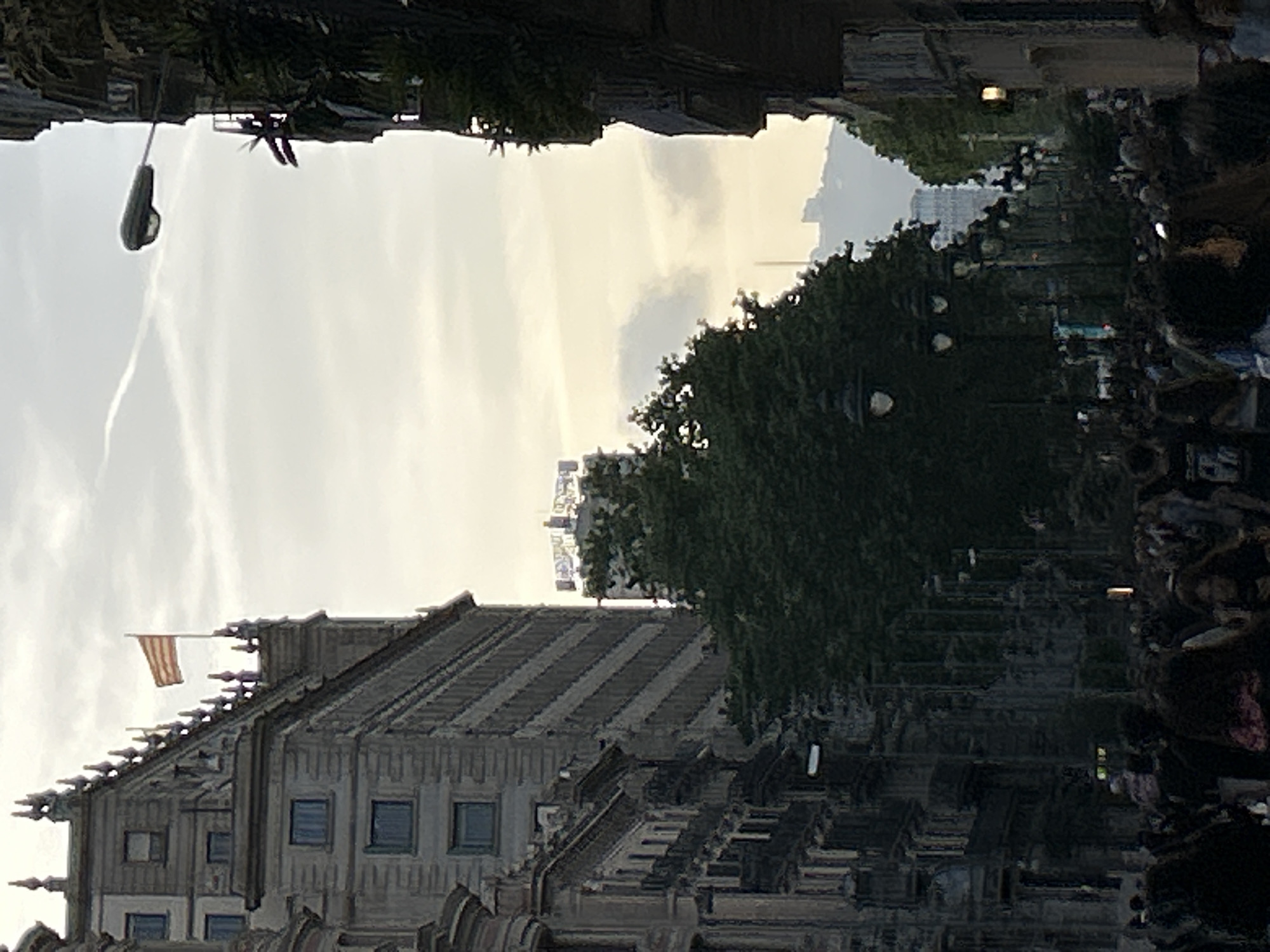 The image, taken in Barcelona, Spain, depicts an urban street scene under a bright, partly cloudy sky. On the left, a grand, ornate historic building dominates the foreground. This multi-story structure is made of stone or concrete, featuring rows of windows and intricate decorative elements, possibly pinnacles or gargoyles, along its roofline. A red and yellow striped flag, consistent with the Catalan or Spanish flag (Senyera), flies prominently from a pole at the top of the building.
To the right, a wide tree-lined street or promenade extends into the distance. Numerous mature green trees form a canopy, partially obscuring a crowd of people gathered or walking along the thoroughfare. Their forms are somewhat indistinct due to the distance and shadows. A modern streetlight fixture is visible high in the sky on the upper left. The sky itself is luminous with soft white clouds and a faint airplane contrail. In the background, beyond the trees and above the historic building, the silhouettes of distant, taller urban buildings can be faintly discerned, reinforcing the city setting. The overall lighting suggests either late afternoon or early evening. There is no visible text in the image.