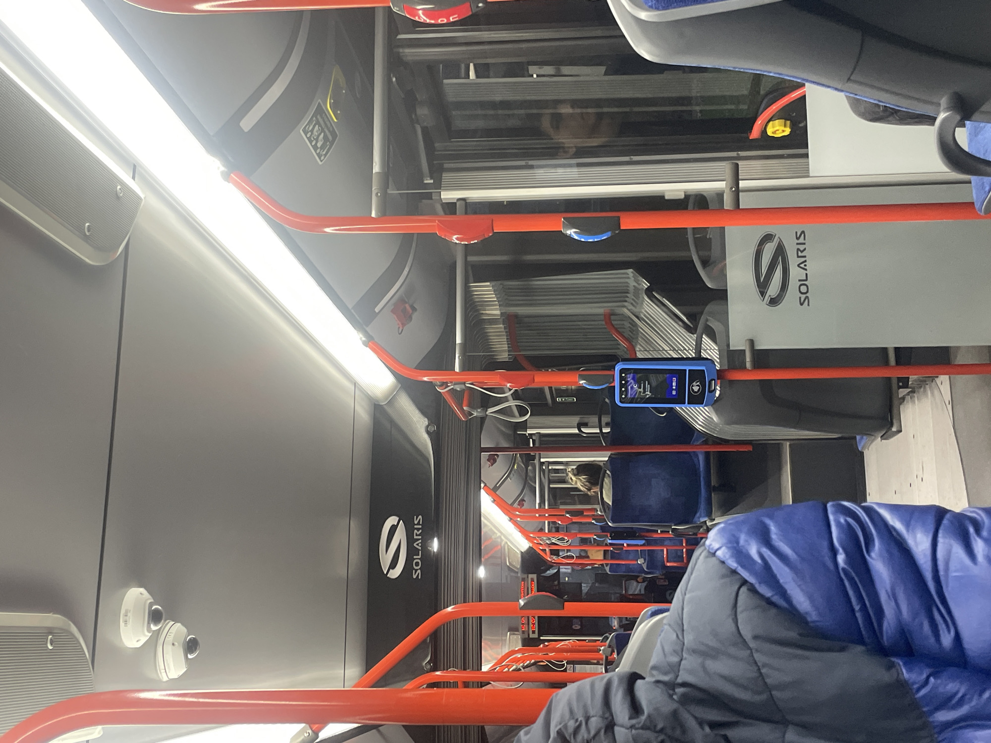 The image shows the interior of a Solaris bus in Sofia, Bulgaria. The bus has orange handrails. There are rows of blue seats visible. Several passengers are seated. A ticket validator is mounted near the top of the bus. The brand name "SOLARIS" is displayed on the bus's side. Two security cameras are mounted on the wall. Fluorescent lights illuminate the bus interior.