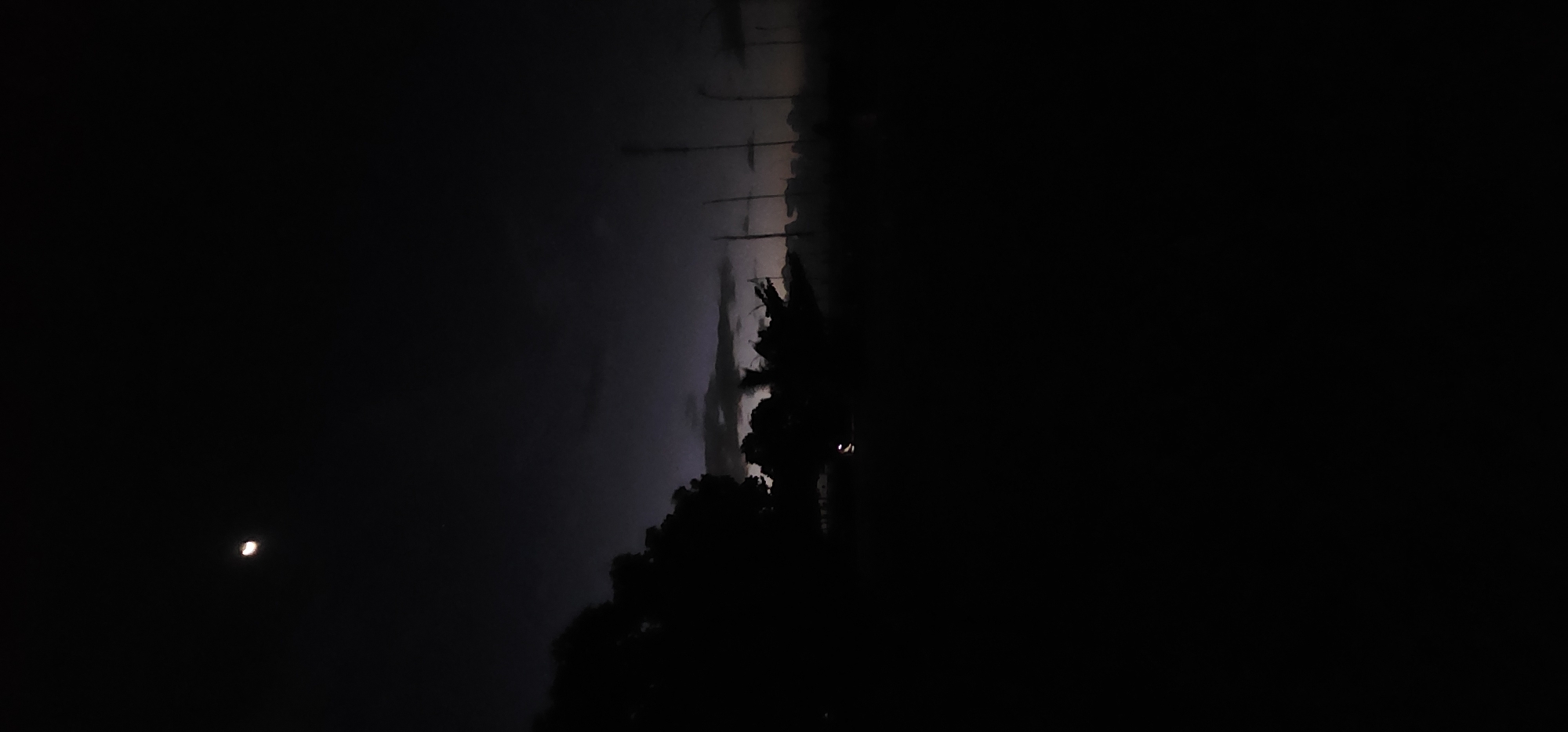 The image shows a dark, night scene. On the left, a crescent moon is visible in the sky. The right side of the image is obscured by darkness, but faint outlines suggest trees and vegetation. A faint glow is seen in the center horizon of the image, indicating a building or structure, possibly in Denpasar, Indonesia. Tall, thin structures that look like poles/towers extend into the background. The overall environment appears to be outdoors, and the lighting is minimal, likely natural.