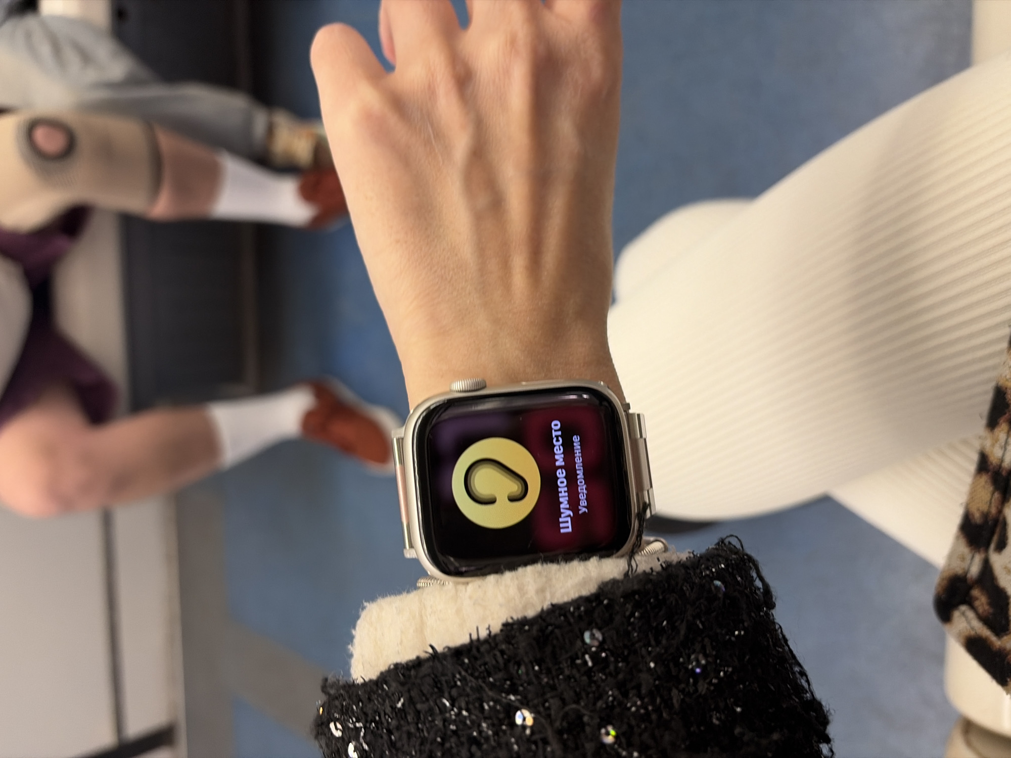 A person's arm is in focus, wearing a silver Apple Watch displaying a notification written in Russian that appears to concern noise levels. The watch rests on a black and white textured fabric. The background is blurred, but shows a person seated with legs crossed, wearing white socks and what appears to be shoes or slippers. A modern white chair is visible in the periphery. Although location is ambiguous, the photograph was taken in Saint Petersburg, Russia.