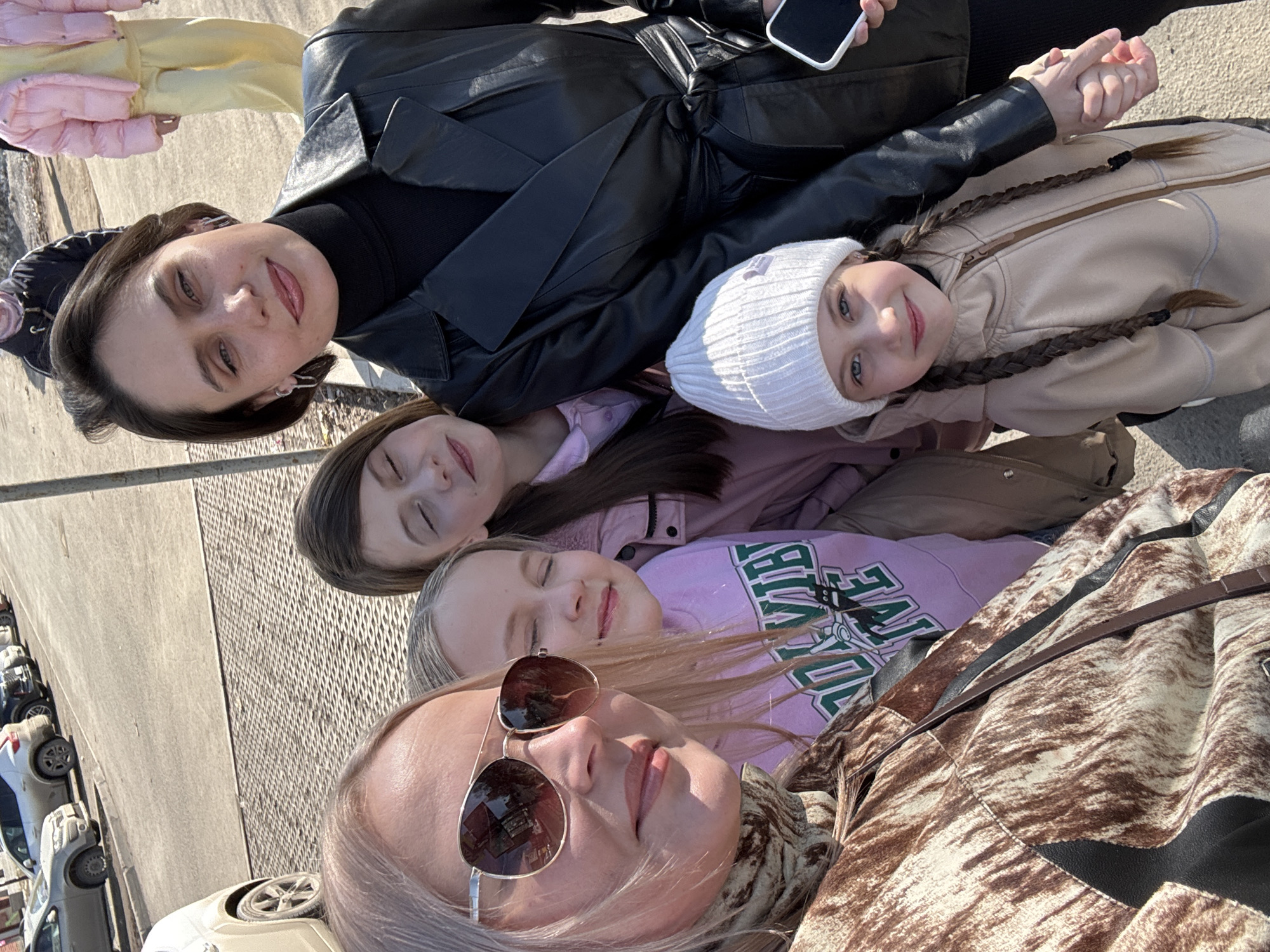 A woman and three girls are lying on a concrete surface in Izhevsk, Russia, presumably outdoors in a public space. The woman closest to the camera is wearing sunglasses and a brown and white patterned jacket. Behind her, a girl in a lavender sweatshirt has her eyes closed. Another girl with closed eyes and brown hair is behind her. A girl with braided hair and a white beanie is next, and behind them is another woman wearing a black turtleneck and a leather jacket, holding a smartphone. The background shows parked cars and a concrete surface.