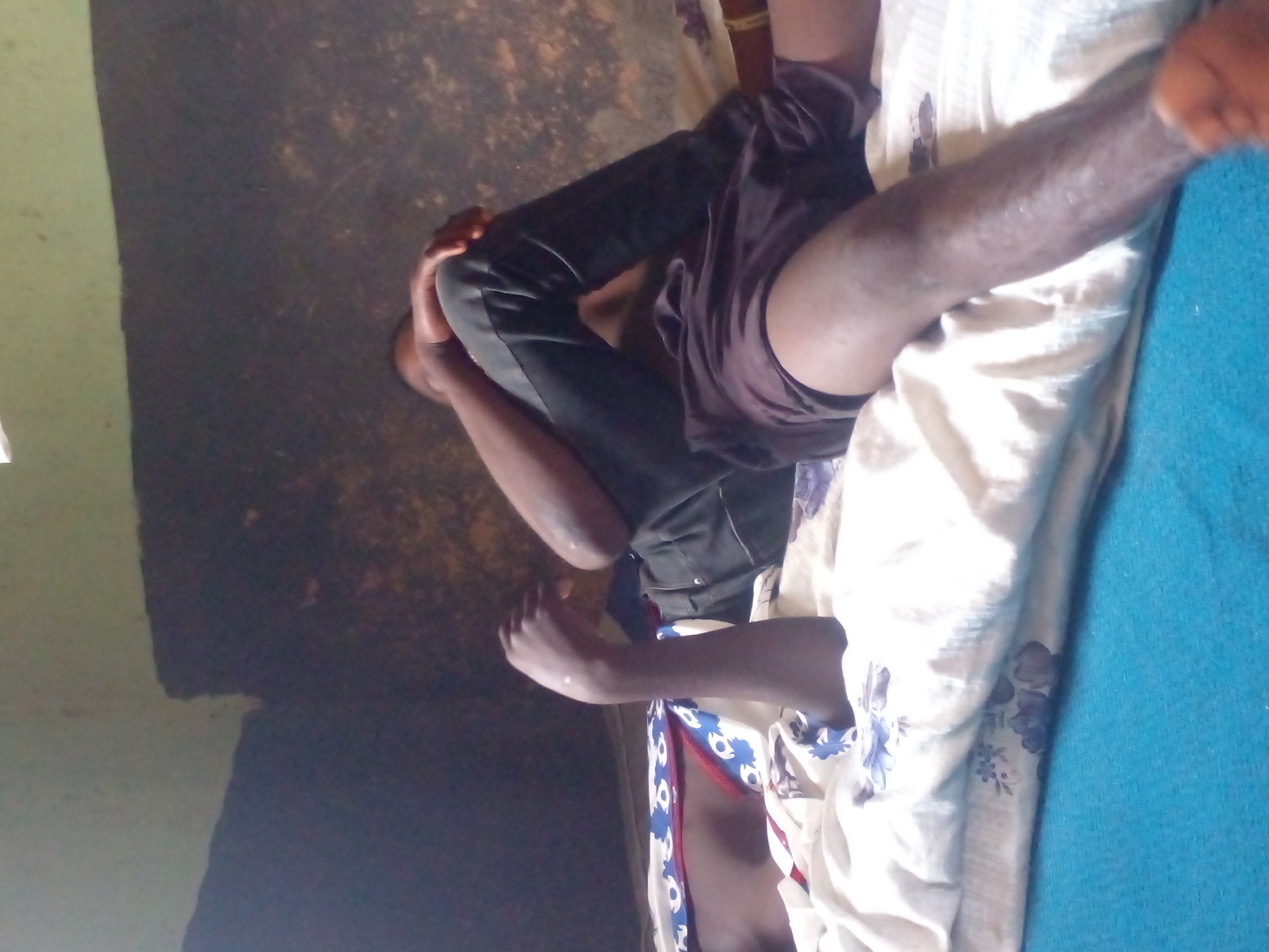 The image shows two individuals lying on a bed. The person in the foreground is lying on their back, with their legs extended. They are wearing dark shorts. The person in the background is lying on their side, facing away from the camera. They are wearing dark pants and their arm is resting on their leg. Both individuals appear to be male and are of dark complexion. The bed is covered with white sheets that have a blue and red floral pattern. The room appears to be sparsely furnished, with a dark, textured wall in the background. The lighting is dim, suggesting it might be indoors. The overall scene depicts a relaxed or resting state.