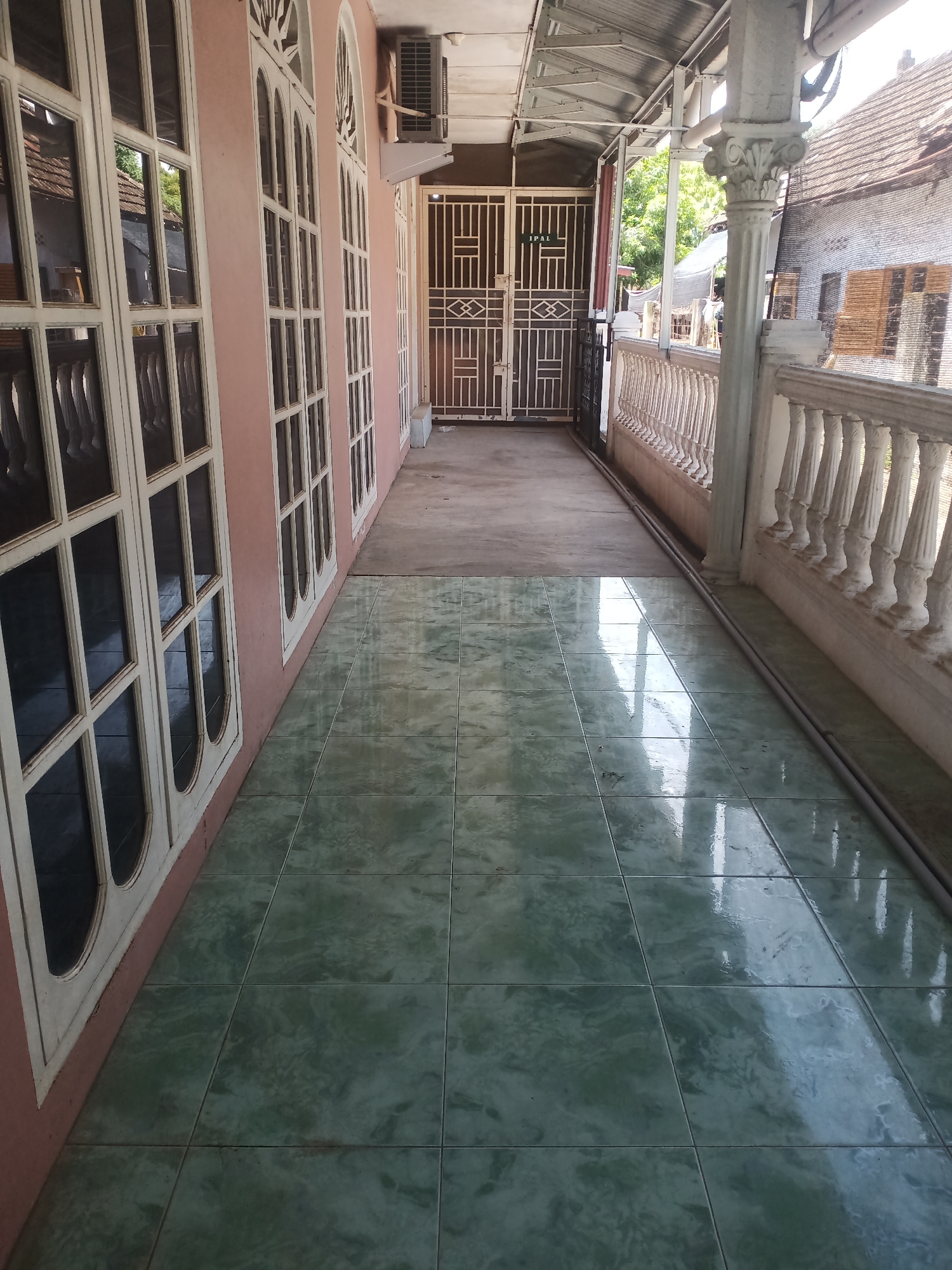 The image depicts the exterior of a building in Lhokseumawe, Indonesia. The foreground shows a tiled floor with a greenish hue, reflecting light and the surrounding structures. To the left, a pinkish wall is visible, adorned with tall, arched windows with white frames and numerous panes. An air conditioning unit is mounted high on this wall. A walkway extends further into the scene, leading to a double gate with a geometric metal design. The walkway is flanked on the right by a white, ornate railing supported by a decorative column. Beyond the railing, a building with a tiled roof and a mesh-covered window is partially visible, suggesting a residential or commercial area. The overall impression is of an outdoor walkway or veranda attached to a building.