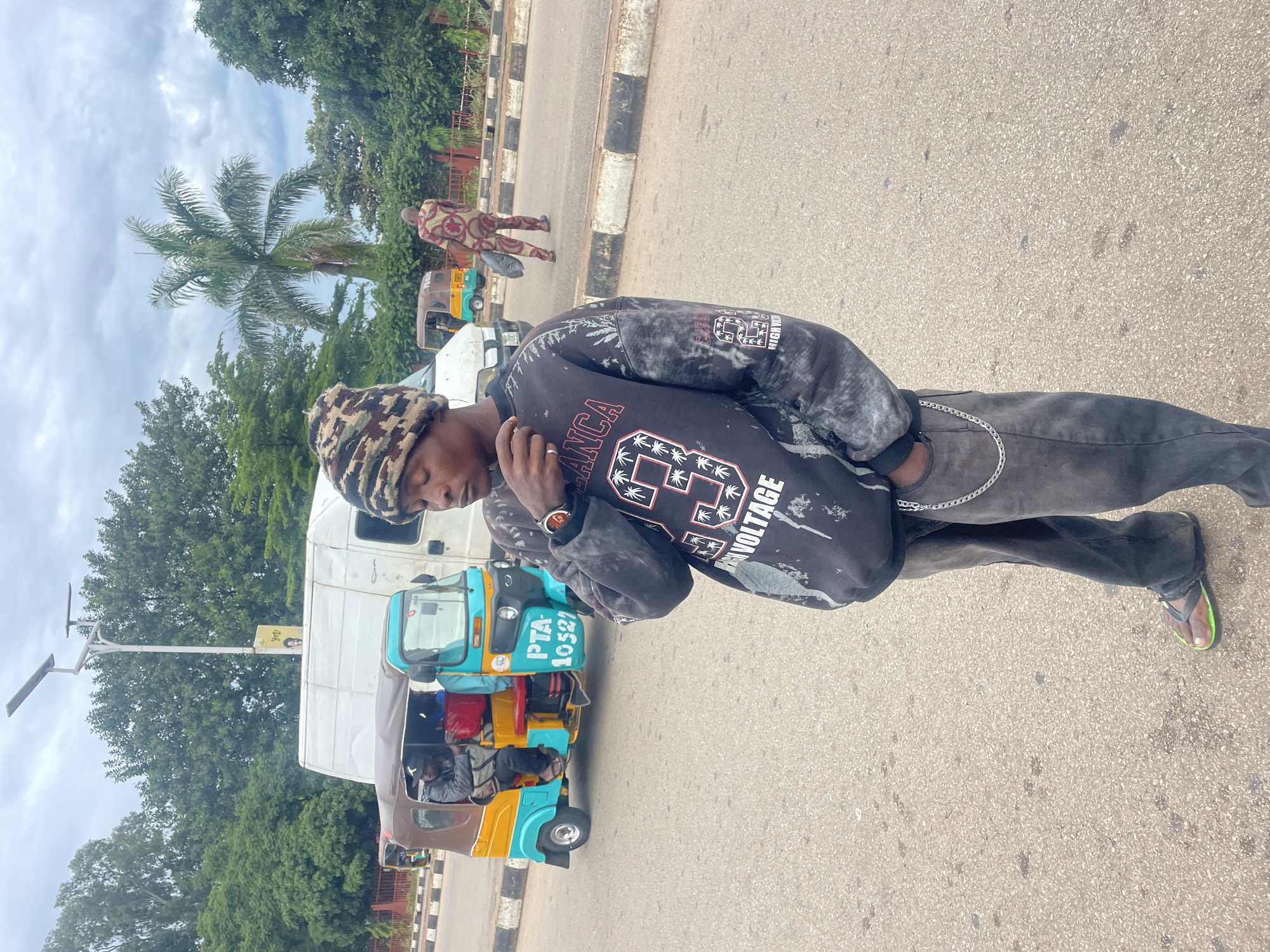 The image depicts a street scene in Barakin Lerawi, Nigeria. In the foreground, a young man stands facing slightly away from the camera, wearing a camouflage beanie, a dark-colored sweatshirt with graphics, and grey pants with a silver chain. He has his right hand near his face. Behind him, a turquoise and yellow auto-rickshaw, a common mode of transport in the region, is visible with passengers inside. A white van is parked behind the auto-rickshaw. Further in the background, a person in a patterned outfit is walking away, carrying a black bag. The environment appears to be an urban setting with trees and a cloudy sky. The road has distinct white and black markings on the side.