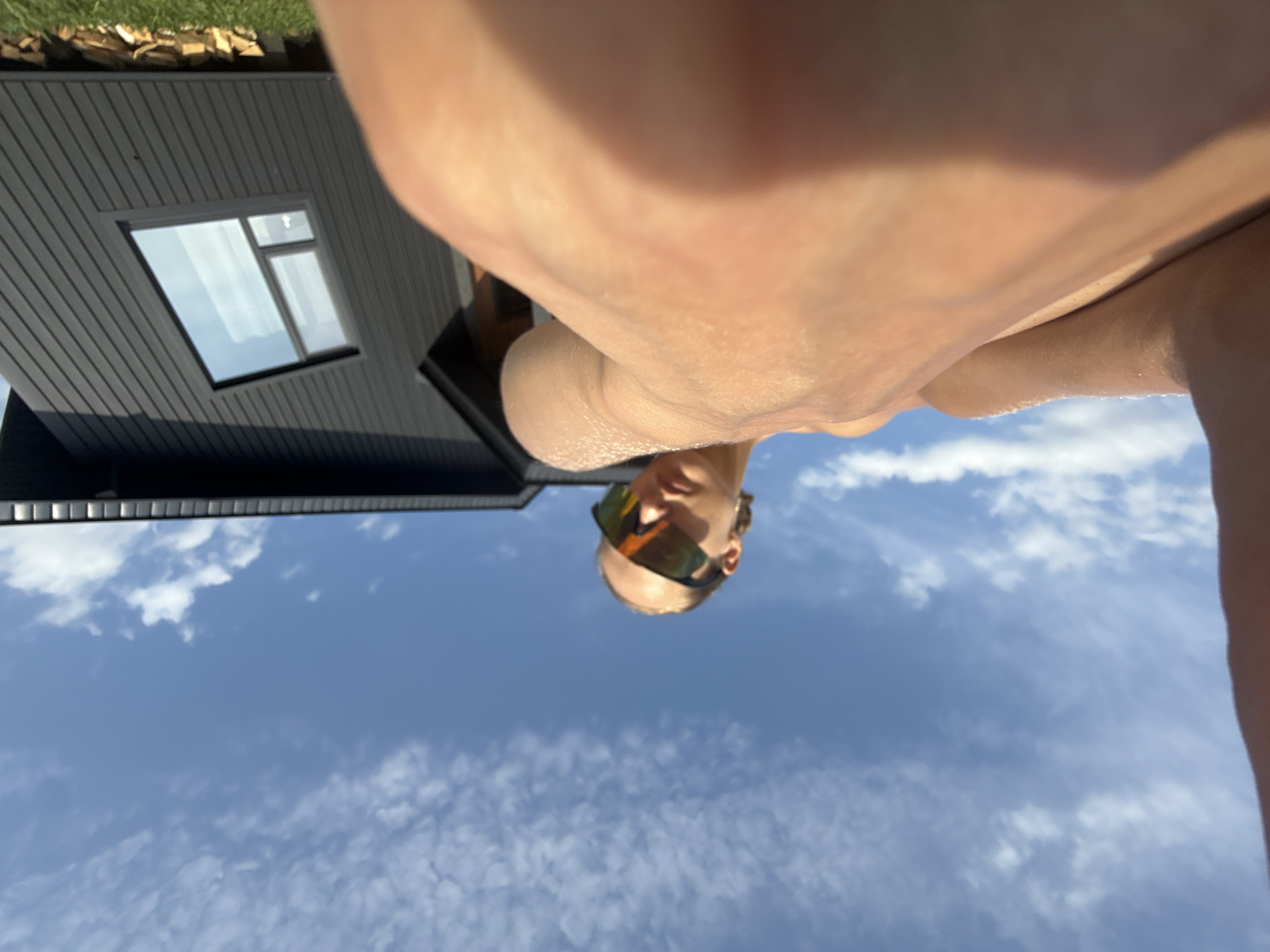 The image depicts a low-angle, close-up perspective of a person wearing reflective sunglasses. The individual's skin is prominently featured in the foreground, with parts of their arm and torso visible. Behind them, a modern, dark-colored building with a distinctive angular window is visible, set against a bright blue sky with scattered clouds. The building appears to be situated in a rural or suburban environment, possibly a private residence, given the presence of stacked firewood and grass in the lower right portion of the frame. The setting suggests an outdoor activity on a sunny day. There is no specific information in the image that allows for identification of the location within the city of Подшивалово, Russia.