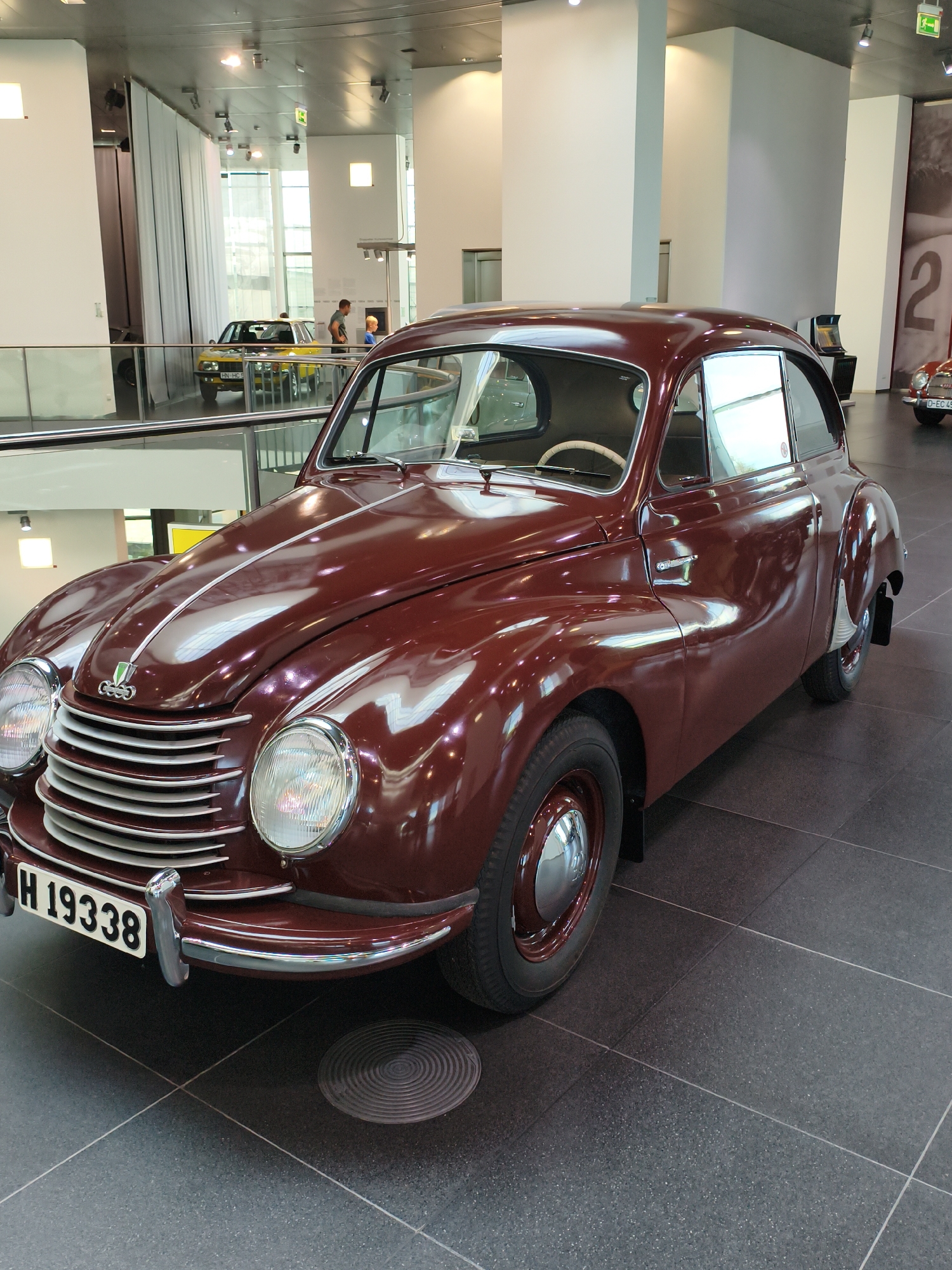 The image shows a maroon vintage car, likely an Auto Union, on display indoors. The car has a rounded design with prominent headlights and a grille. A license plate with "H 19338" is visible on the front. The car is parked on a dark tiled floor. In the background, there are white pillars, a light-colored wall with a large "2" graphic, and other vehicles, including a yellow car. The environment appears to be a museum or exhibition hall, consistent with the Audi Forum in Ingolstadt, Germany, which houses the Audi museum mobile. There are no people clearly interacting with the maroon car in the foreground.