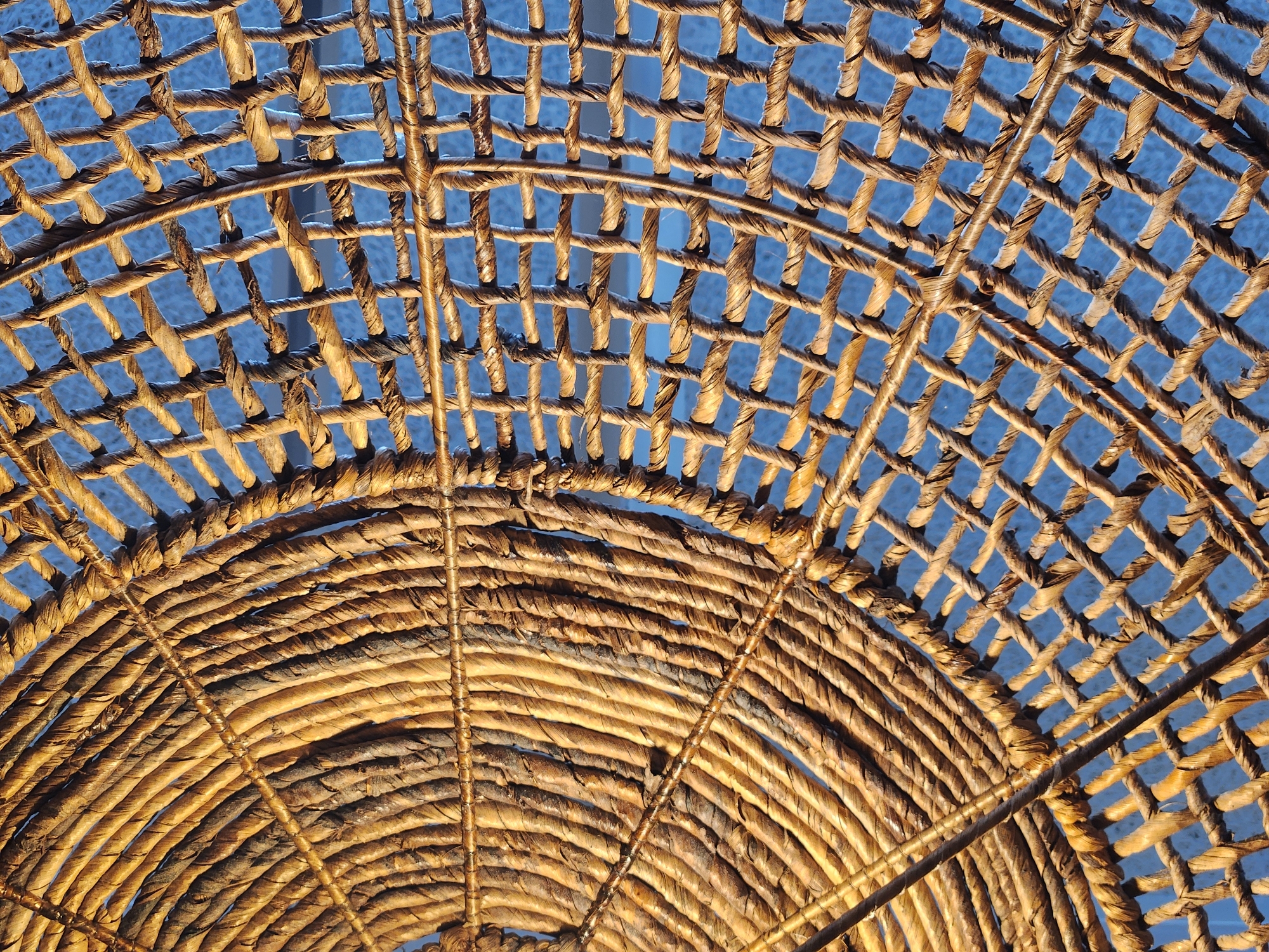 The image is a close-up, overhead shot of woven natural fibers, likely rattan or straw, against a bright blue background, possibly the sky. The woven material forms two distinct patterns: one section on the left exhibits concentric, radiating lines of tightly twisted fibers, resembling spokes of a wheel. The right side features a grid-like pattern of woven strands, creating square or rectangular openings through which the blue background is visible. The lighting suggests it is daytime, casting warm tones on the fibers. The texture is rough and organic, with variations in color and thickness of the individual strands. There are no people, animals, or discernable landmarks visible. The scene is abstract and focused solely on the texture and pattern of the woven material. The location within Maitland, United States of America, cannot be determined from the visual information provided in the image.