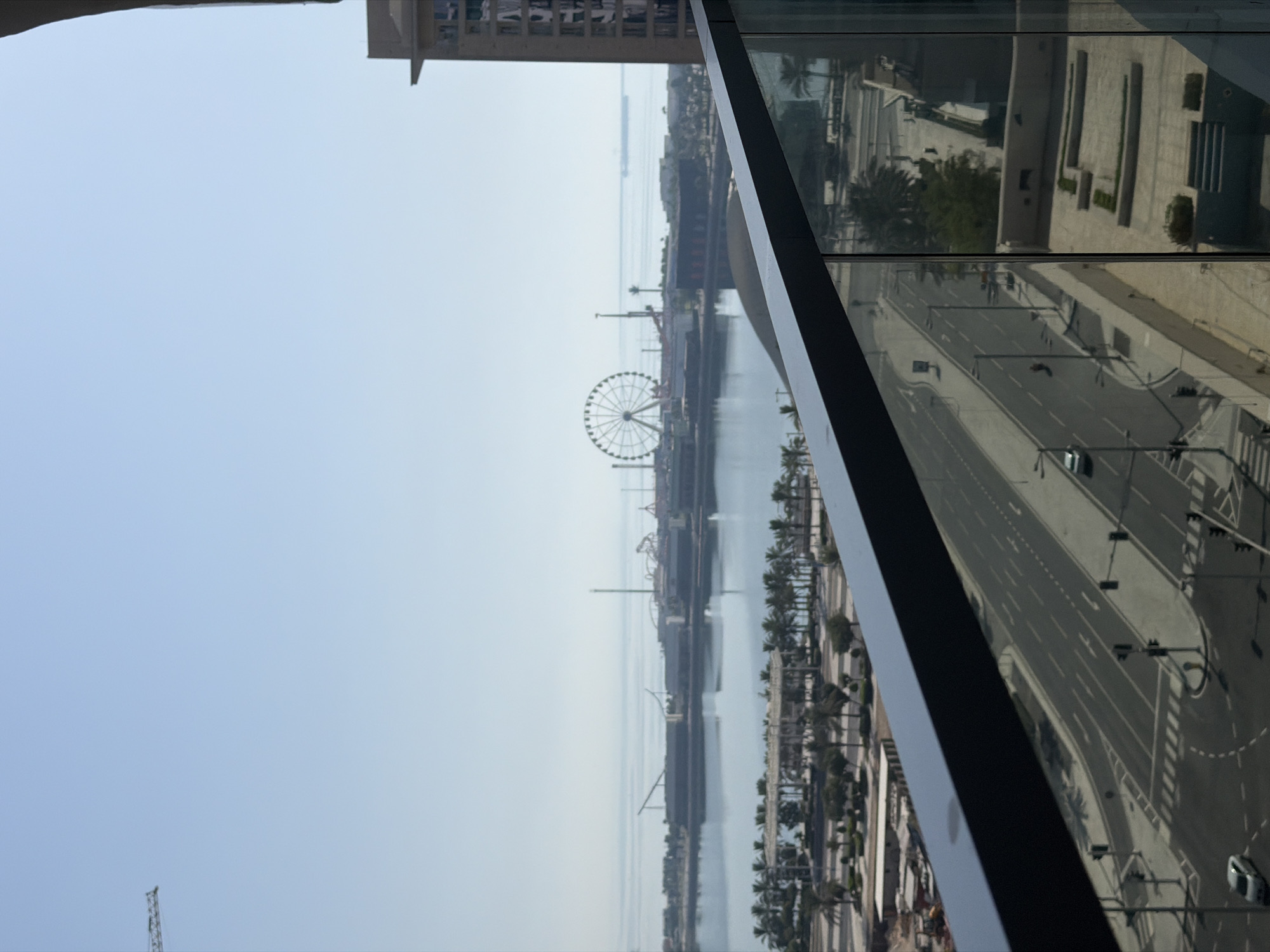 The image captures a wide-angle view from a high vantage point, likely a balcony, looking out over a coastal city. In the foreground, a dark, angled railing of the balcony cuts across the lower portion of the frame, with a reflective glass surface to its right. Below the balcony, a multi-lane road with traffic lights and lane markings is visible, with a few cars present. Further into the mid-ground, a body of water, possibly a bay or harbor, separates the foreground from a distant shoreline. Along this shoreline, an amusement park with roller coasters and a prominent Ferris wheel is situated. Palm trees are visible in a landscaped area between the road and the water. The background is dominated by a clear, pale blue sky, with a hint of the horizon where the water meets the sky. A construction crane is visible in the upper left quadrant of the image, and a tall building with visible windows stands to the right. The overall impression is one of a modern, sunny urban environment by the sea.