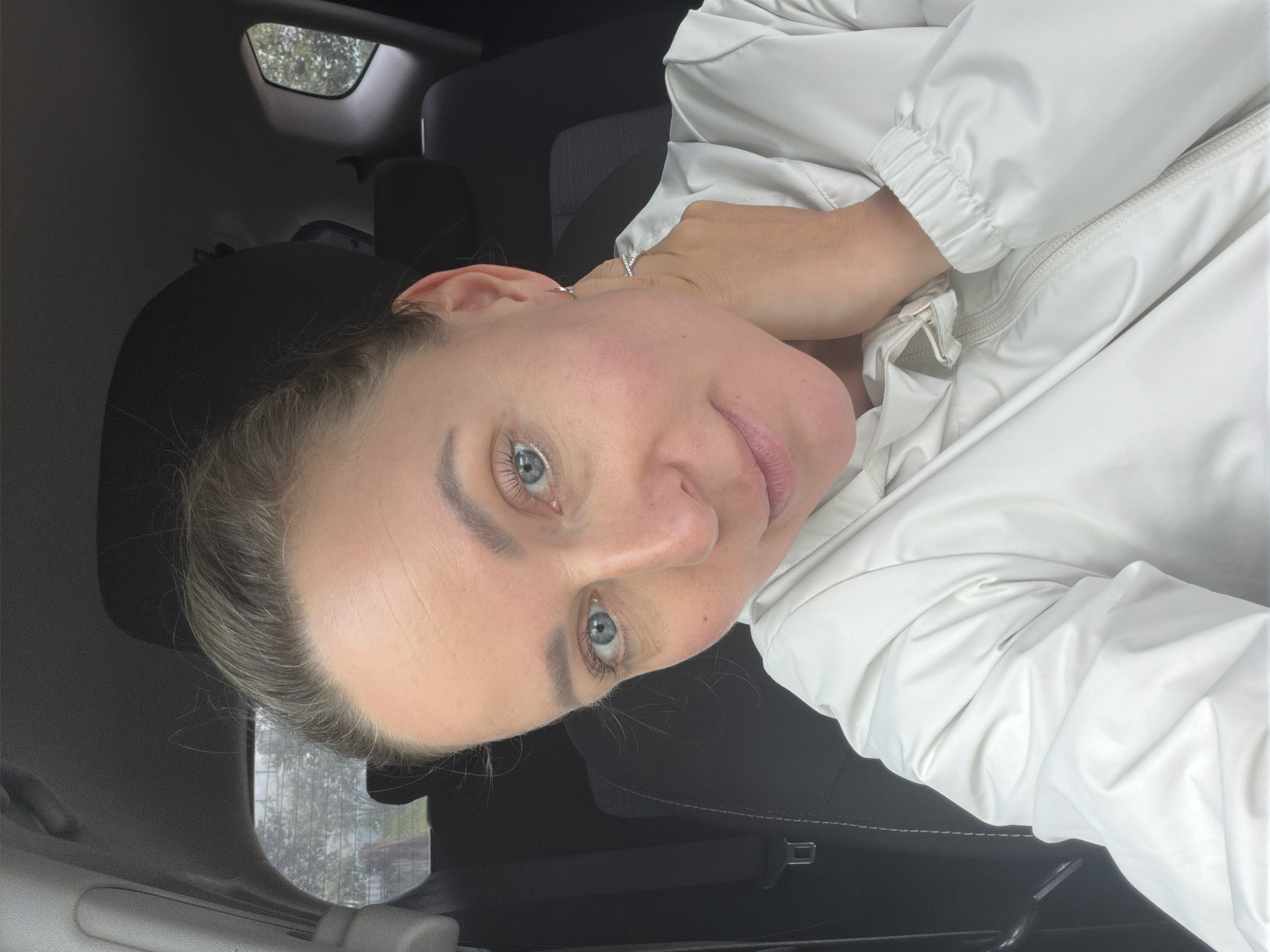 A woman with blonde hair pulled back and blue eyes is sitting in the driver's seat of a car. She is wearing a white jacket. Her right hand is touching her neck. The interior of the car is visible, including the headrest and seatbelt. Outside the car, trees and a cloudy sky can be seen through the windows.