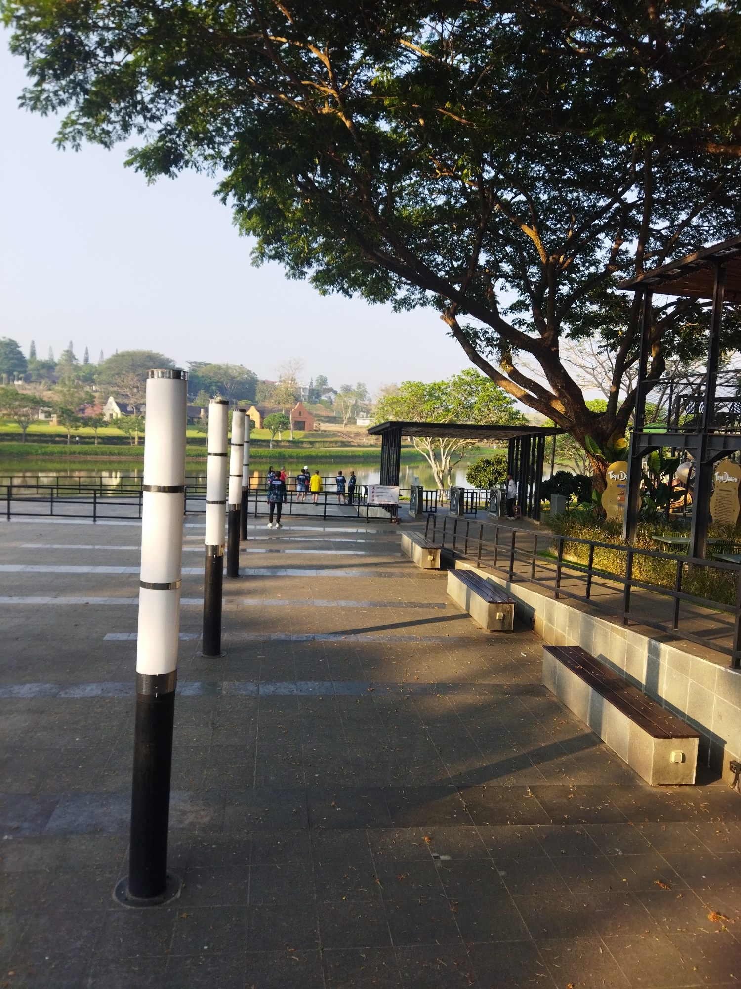 This image depicts an outdoor scene, likely a park or recreational area, with a focus on a paved walkway lined with modern lampposts and benches. In the background, a calm body of water, possibly a lake or reservoir, is visible, reflecting the surrounding landscape. Across the water, a charming village-like settlement with quaint houses and churches can be seen, nestled among lush green trees and hills. The overall atmosphere is serene and tranquil, suggesting a peaceful day in a picturesque setting. The location is identified as Semarang, Indonesia.