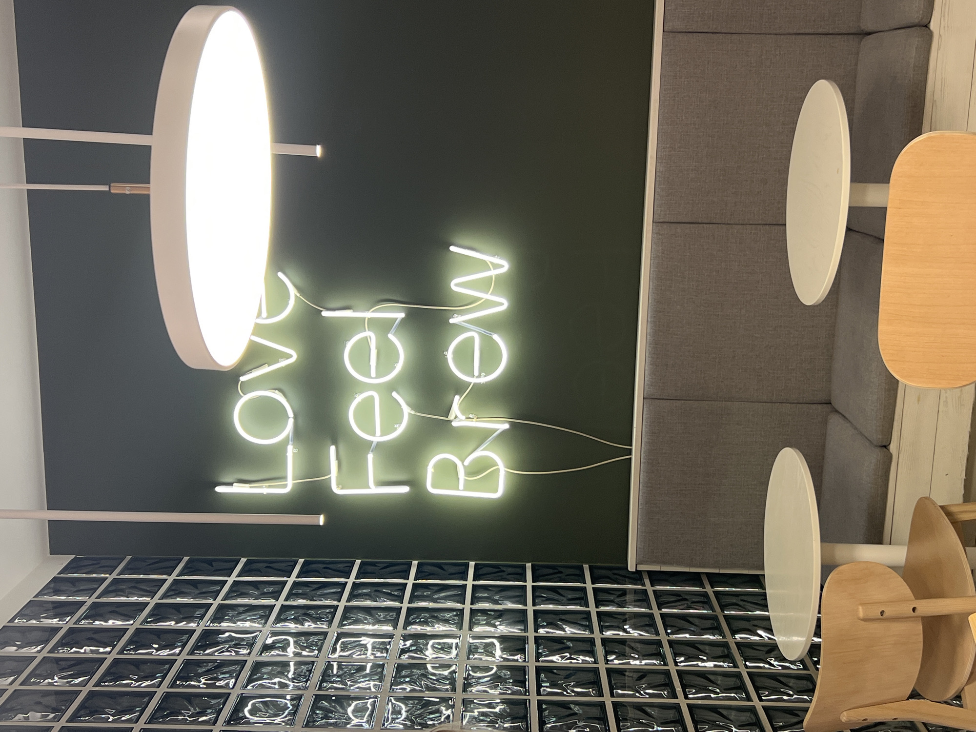 A brightly lit interior in Novosibirsk, Russia, featuring a dark green wall adorned with a neon sign that reads "LOVE Feel Brew." Below the wall is a decorative black grid-like surface. To the right, there's a gray upholstered booth with two attached circular tables, one white and one light brown. Adjacent to the table are two light wooden chairs. An off-center, large, circular light fixture dominates the left side of the image.