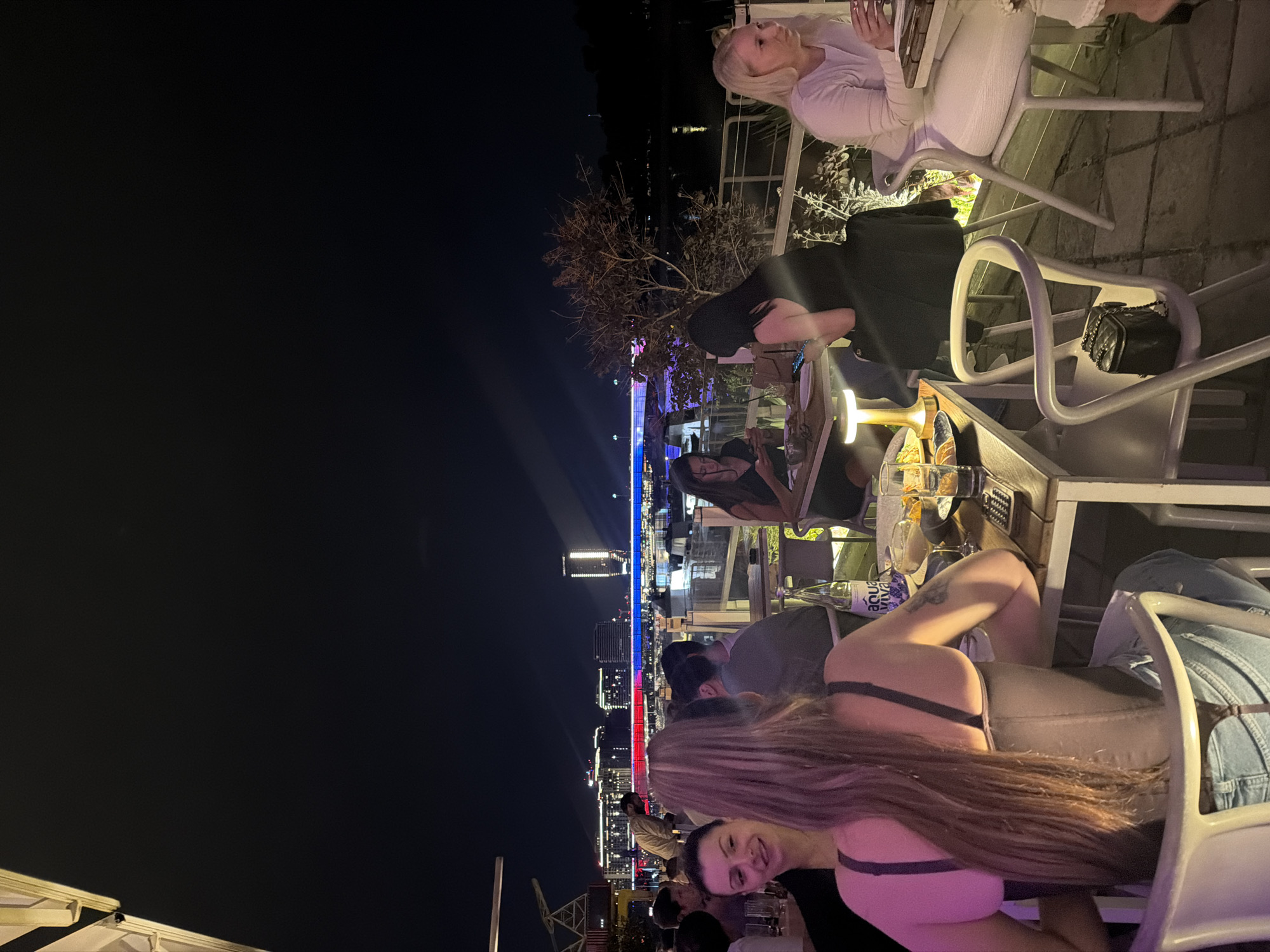A nighttime outdoor social scene shows multiple people seated at tables on a rooftop patio. In the background, the lit skyline of Belgrade, Serbia, is visible. At the foreground, a woman with long brown hair is seated. Other people are seen in the midground. The furniture is a light color, and the tables hold drinks and food. The night sky is dark.