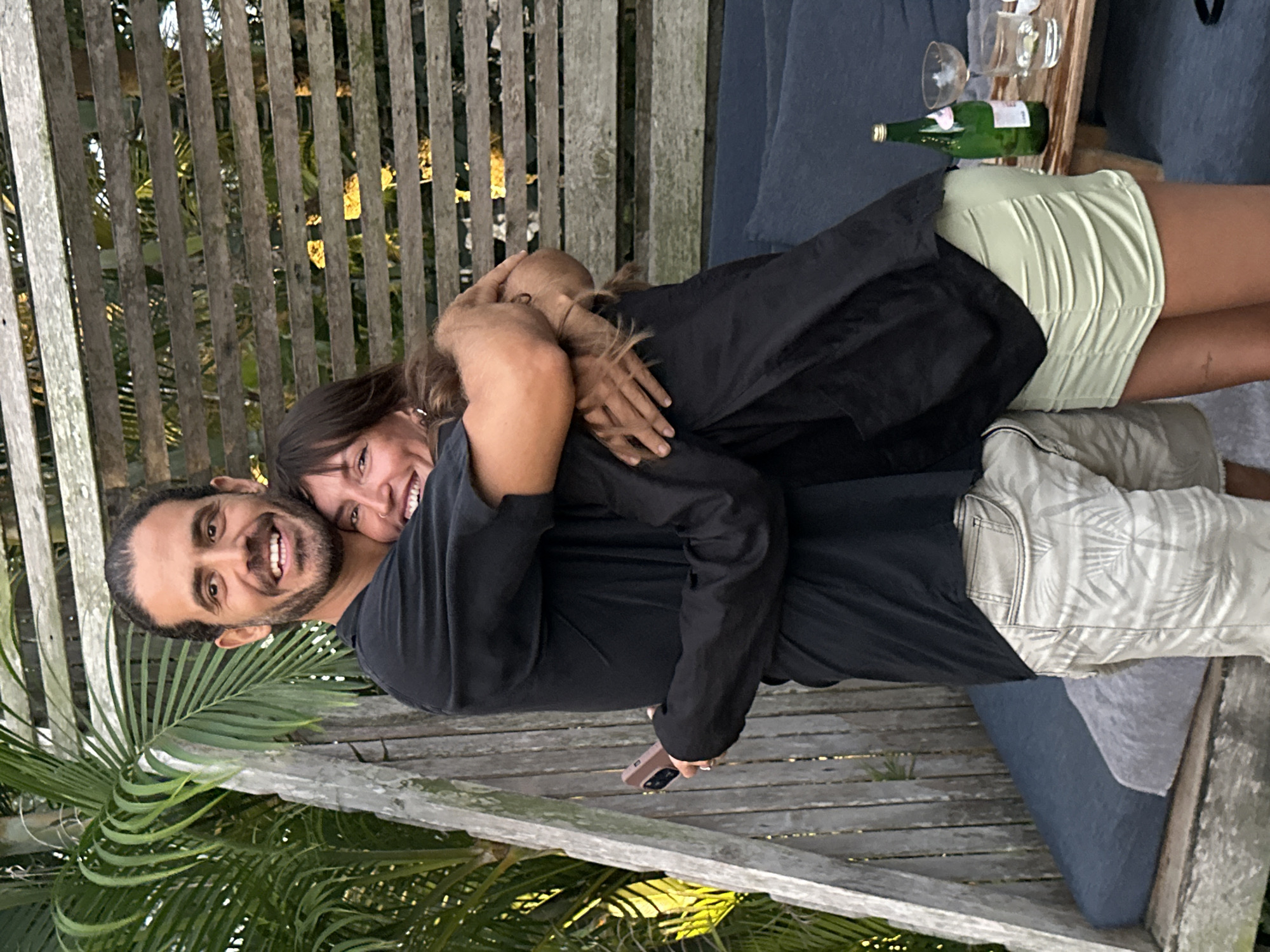 The image shows a man and a woman embracing on a wooden bench outdoors, with a lush green palm frond in the foreground. The man, with dark hair and a beard, is smiling broadly and looking towards the camera. He is wearing a black t-shirt and patterned shorts. The woman, with long brown hair, is nestled into his shoulder, her face partially obscured as she smiles. She is wearing a black top and a light green skirt. Behind them, a wooden structure with vertical slats serves as a backdrop, and a glimpse of a blue cushioned bench is visible. On a wooden surface behind them, a green bottle of liquid, a wine glass, and another glass are placed. The lighting suggests it is late afternoon or early evening, with a warm glow filtering through the foliage. The overall mood appears affectionate and relaxed, set in a tropical or semi-tropical outdoor environment. The image is likely taken in Kuta Selatan, Indonesia, given the palm fronds and general ambiance. No visible text is present in the image.